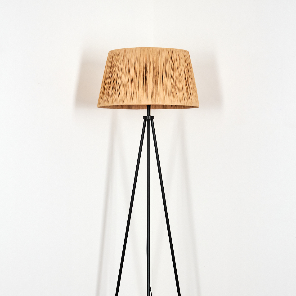 Eine hohe Stehlampe mit gewebtem beigem Schirm und schwarzen Dreibeinbeinen steht vor einer weißen Wand.