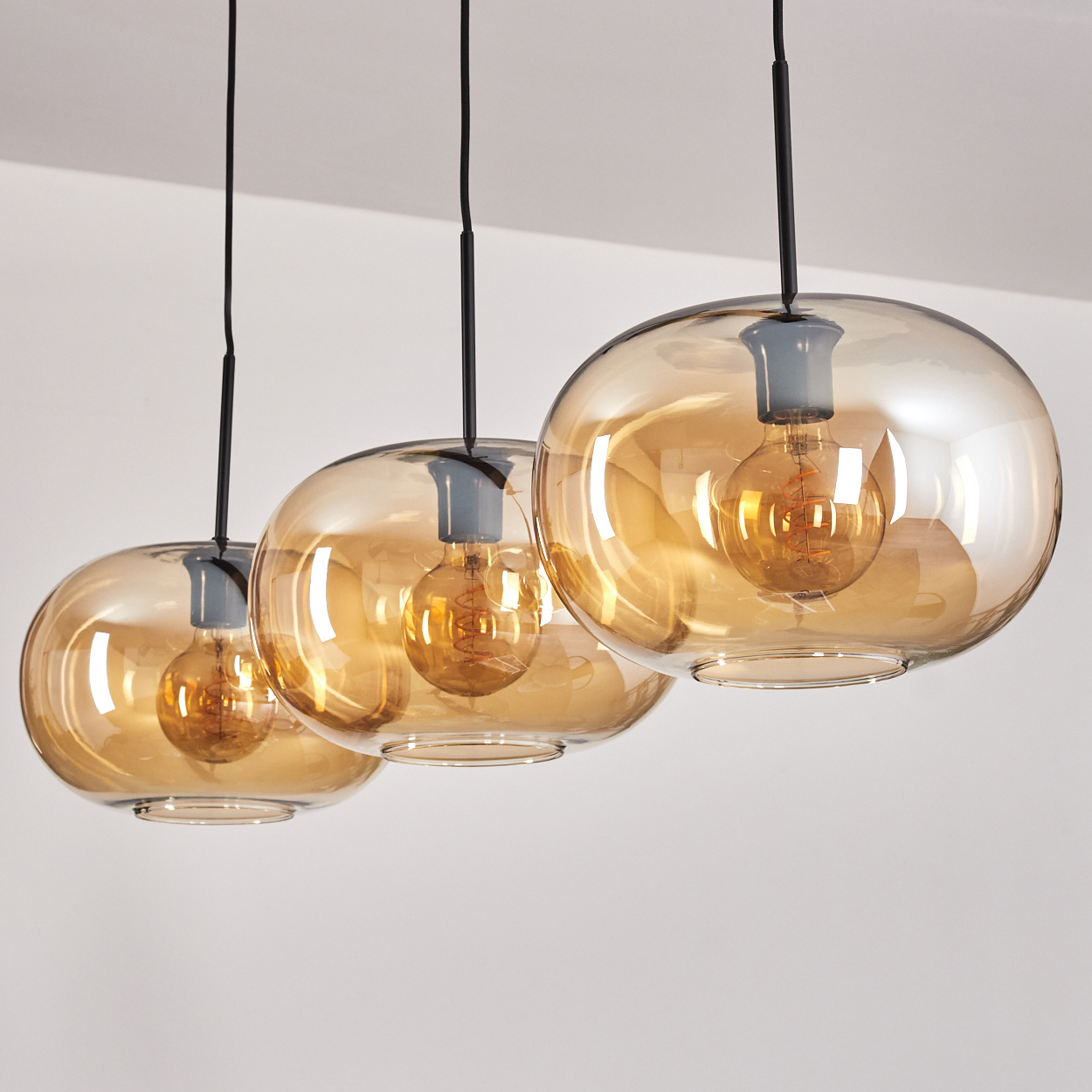 Drei bernsteinfarbene Glaspendelleuchten hängen an schwarzen Kabeln, jede mit Edison-Birne.