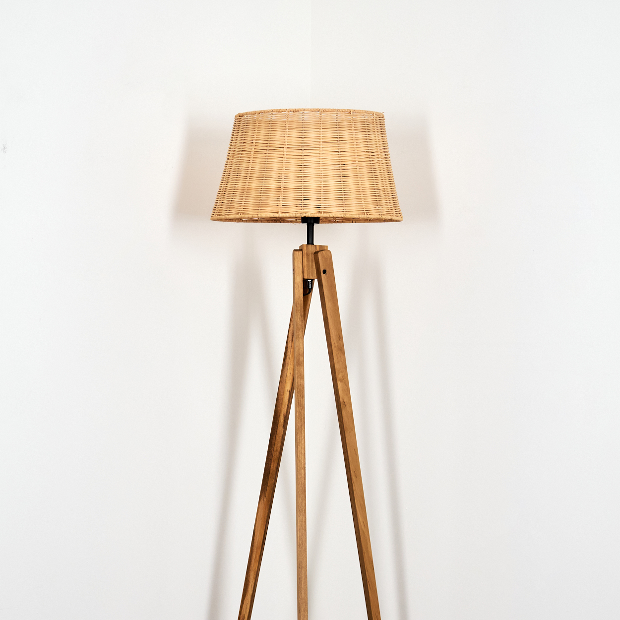 Ein Stehlampen-Stativ aus Holz mit einem geflochtenen Lampenschirm vor einer weißen Wand.
