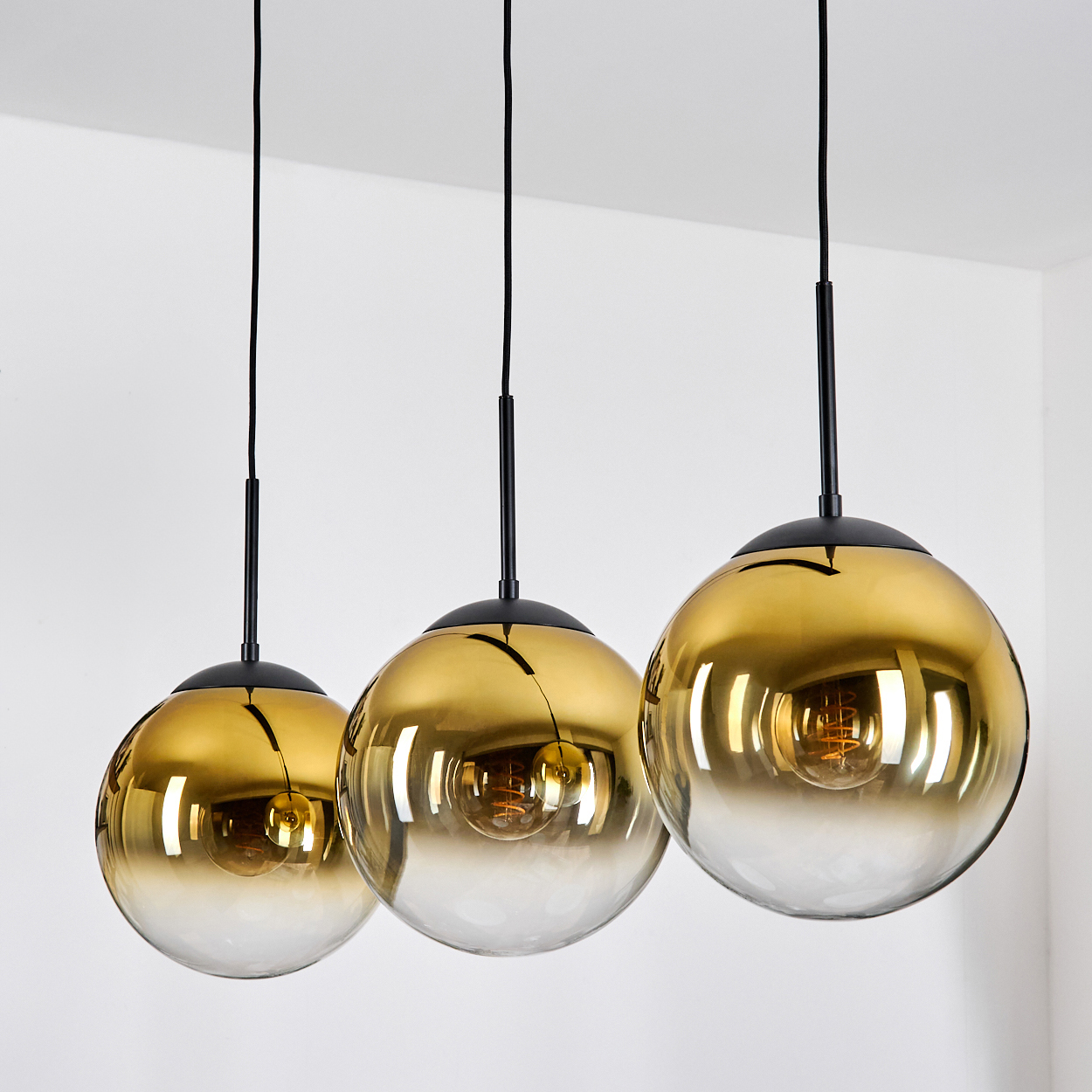 Drei goldene Pendelleuchten mit Glaskugeln hängen an schwarzen Kabeln.