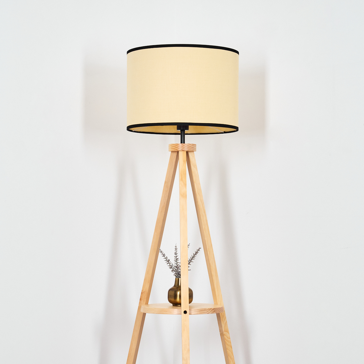 Eine Stehlampe mit hellem Lampenschirm und Holzstativ, daneben ein kleiner goldener Topf mit Zweigen.
