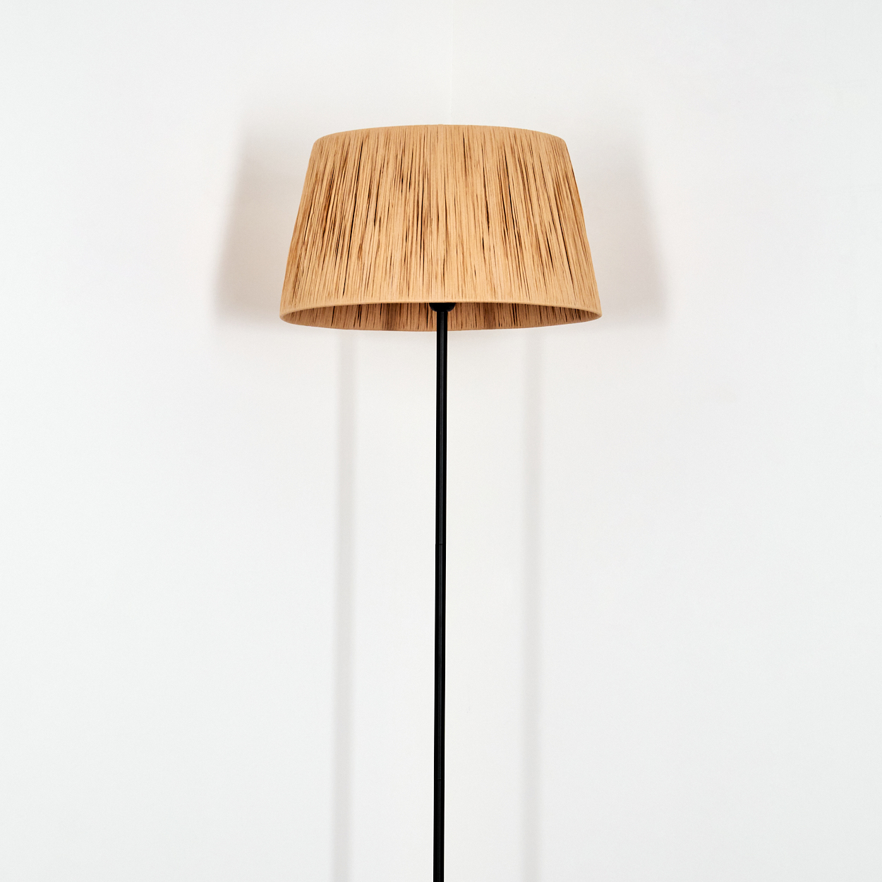 Eine Stehlampe mit einem geflochtenen Lampenschirm aus beigem Stoff vor einer weißen Wand.