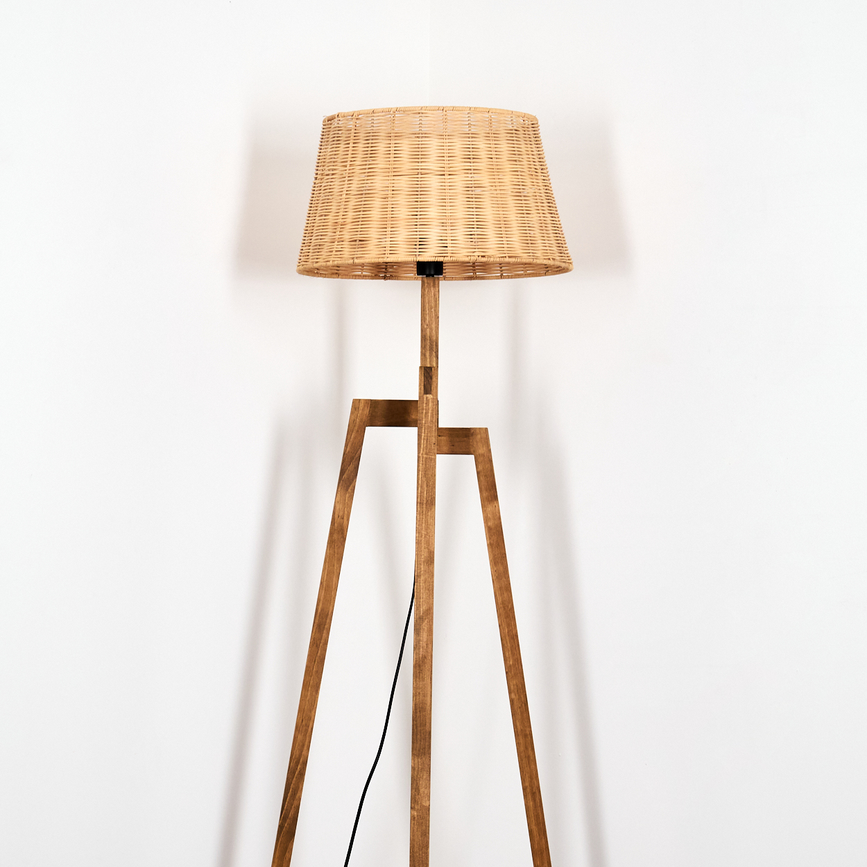 Ein Stehlampenfuß aus Holz mit einem geflochtenen Lampenschirm steht vor einer weißen Wand.