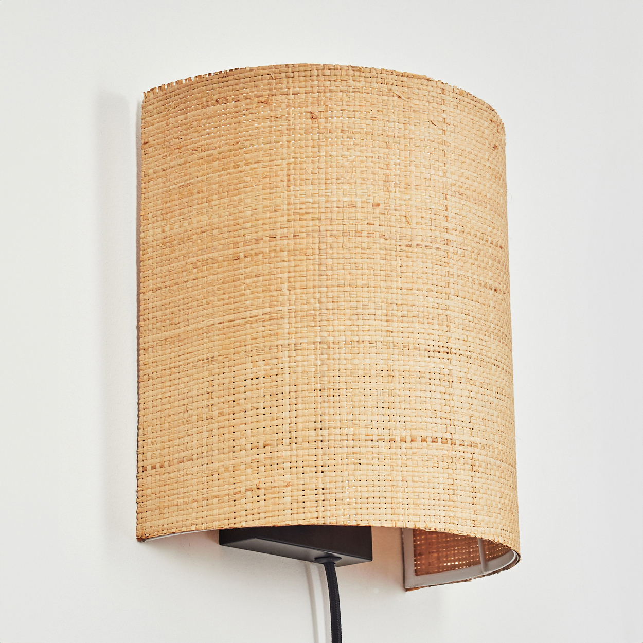 Braune Rattan-Wandleuchte mit schwarzem Sockel und Kabel an weißer Wand.