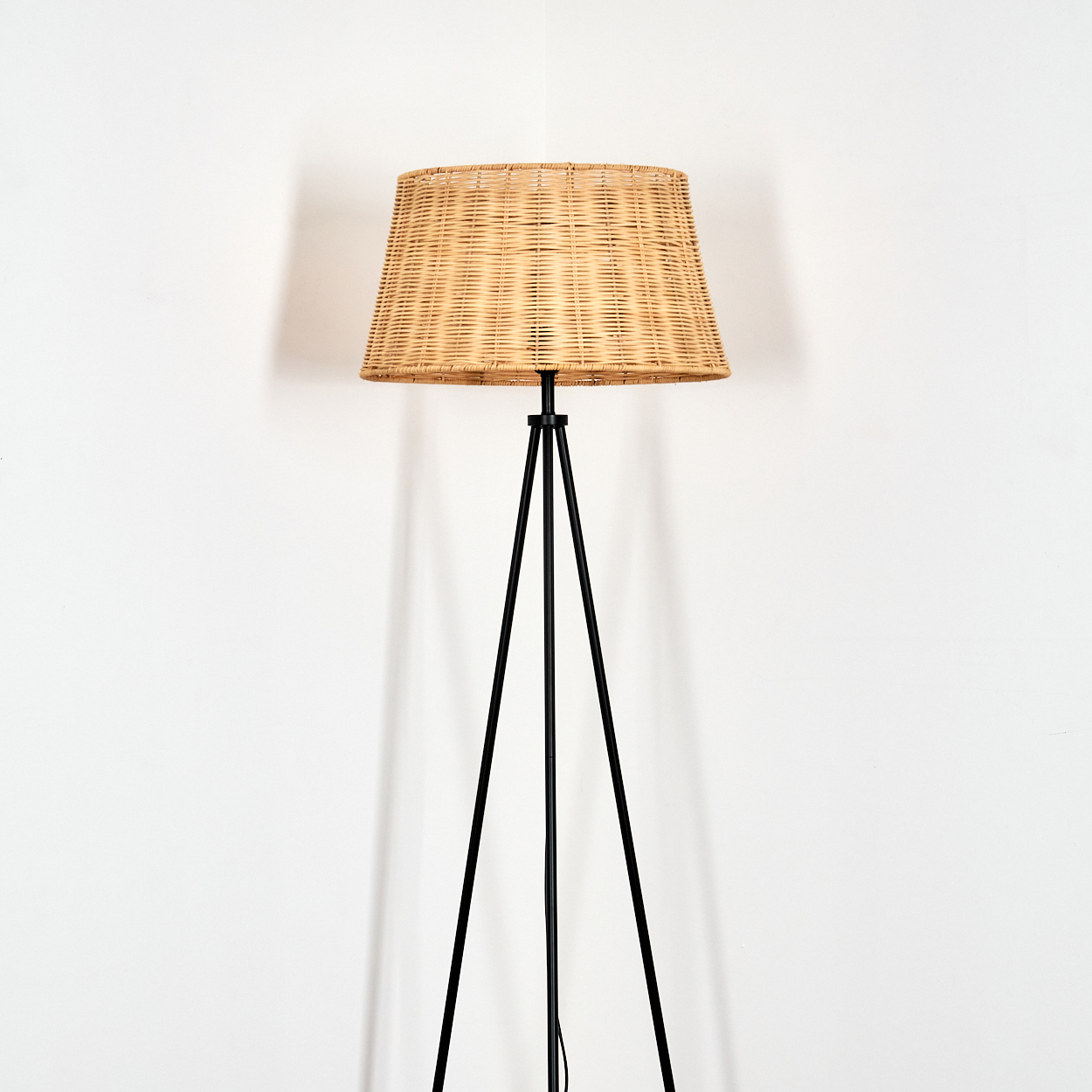 Ein Stehlampe mit einem geflochtenen Lampenschirm und einem schwarzen Stativ steht vor einer weißen Wand.