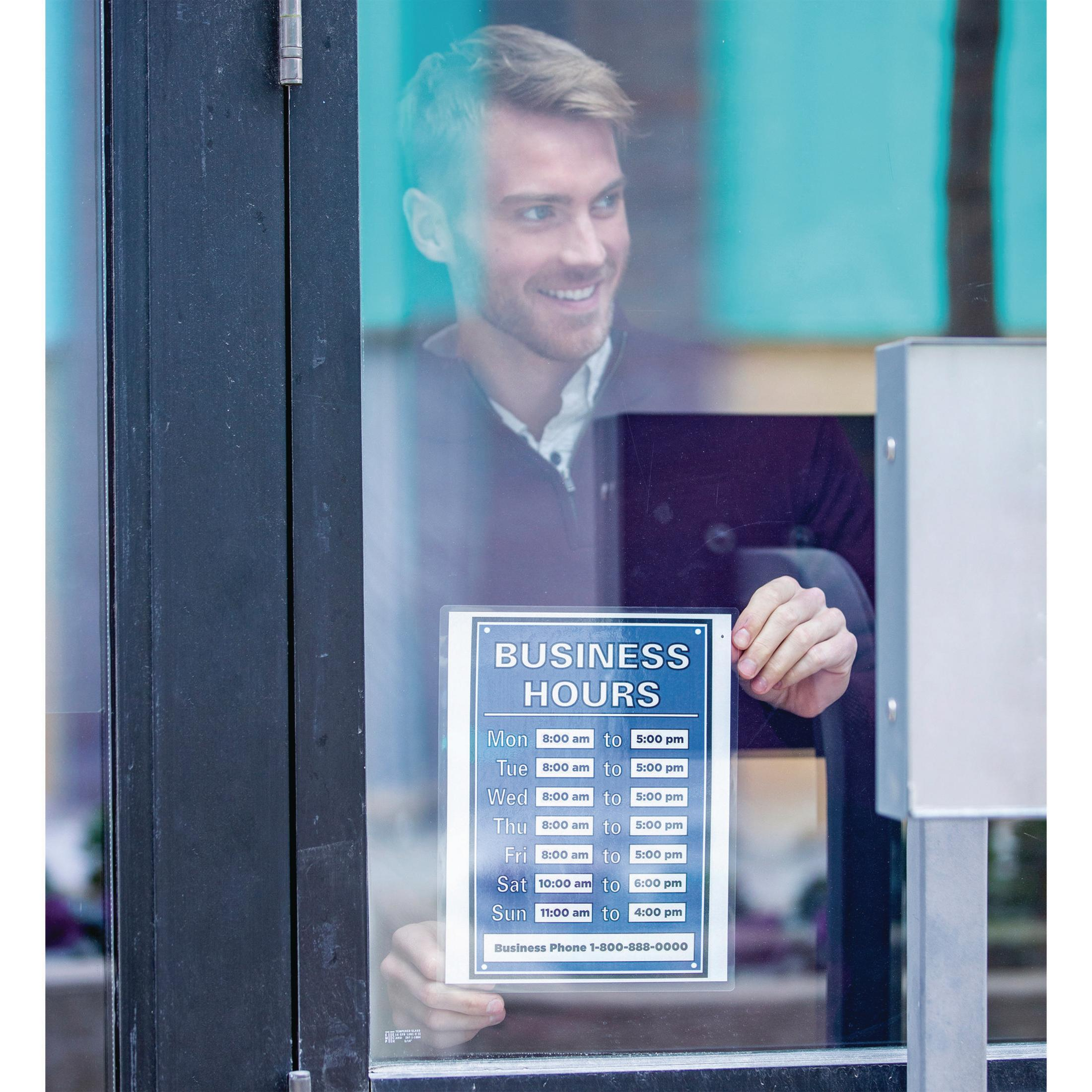 Homme tenant une enseigne avec horaires d'ouverture et numéro de téléphone sur une porte vitrée.