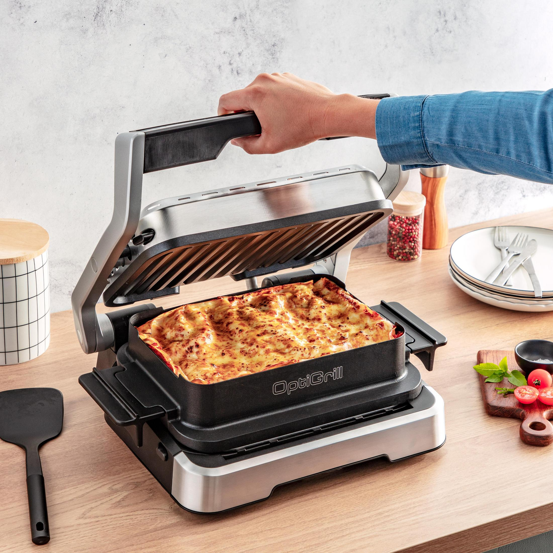 Eine Person öffnet einen OptiGrill mit Lasagne darin. Auf dem Tisch liegen Gewürze und Geschirr.