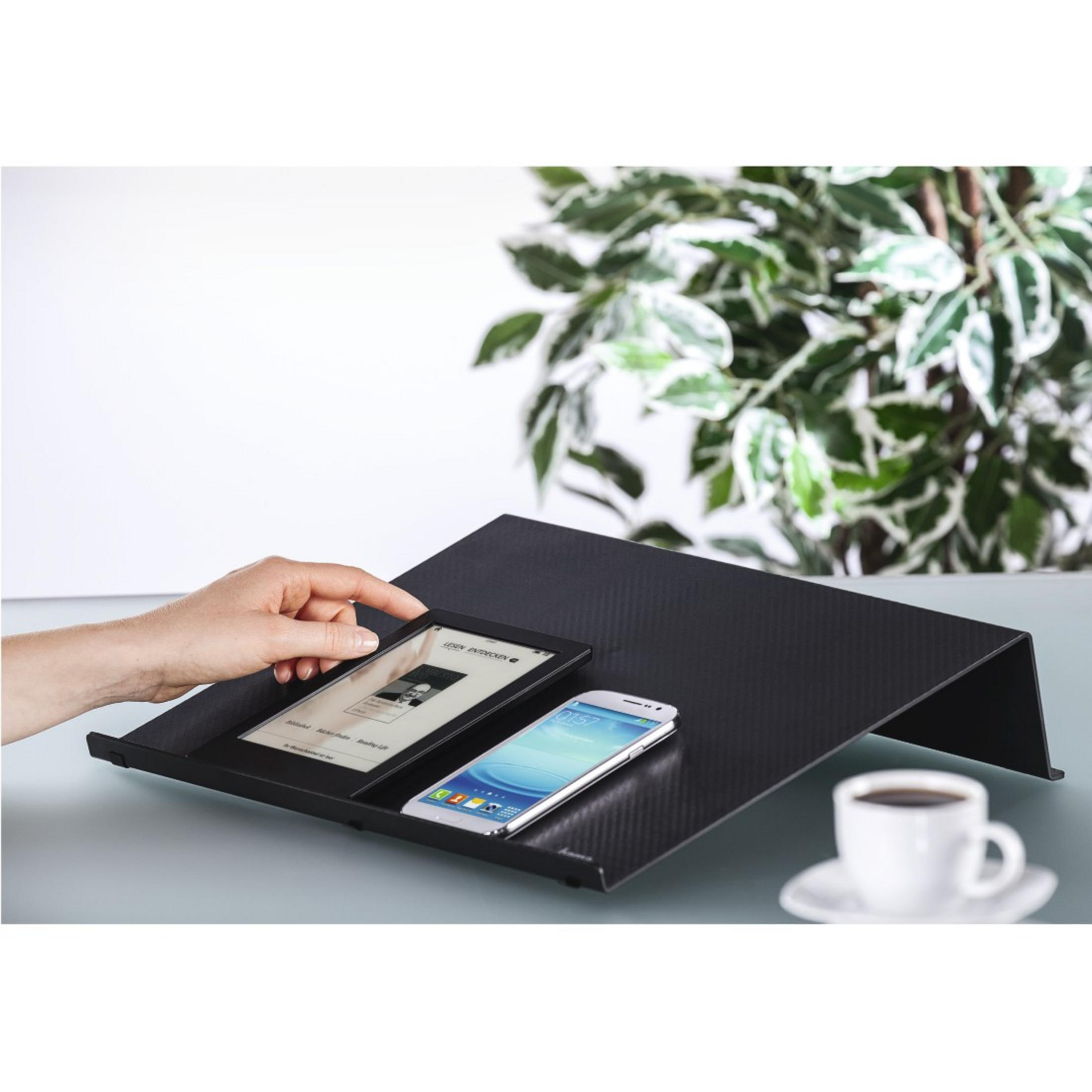 Eine Hand berührt ein Tablet neben einem Smartphone auf einem schwarzen Ständer. Kaffeetasse im Vordergrund.