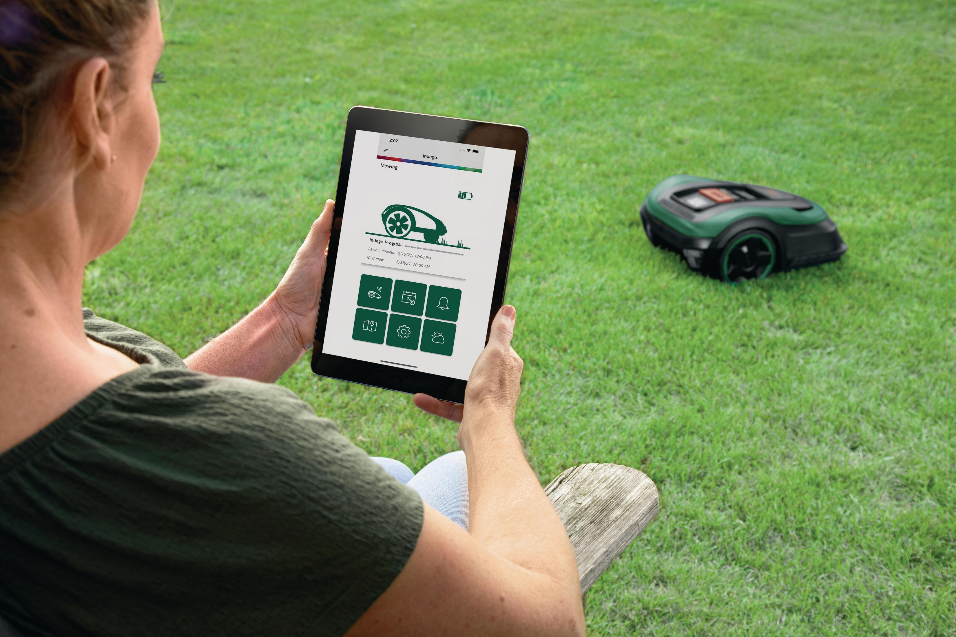 Eine Frau hält ein Tablet mit einer Rasenmäher-App. Ein Mähroboter ist im Gras.
