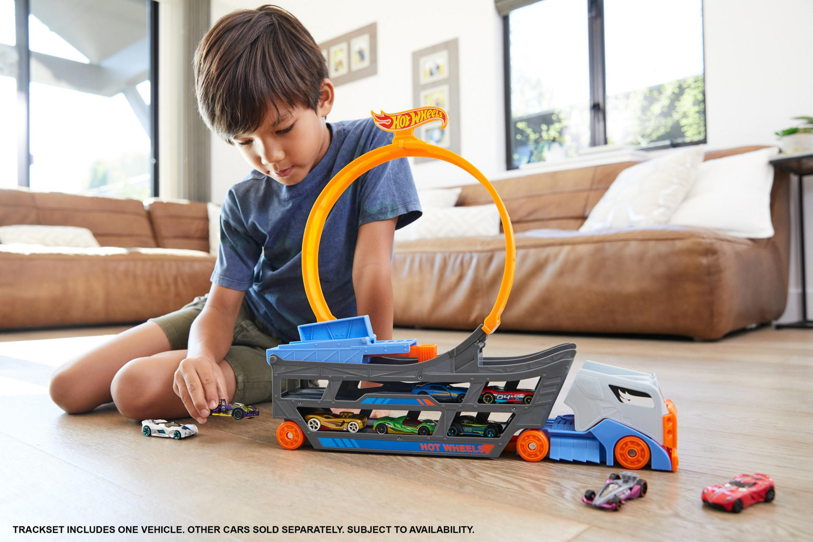 Un garçon joue avec un porte-voitures Hot Wheels et des voitures par terre.