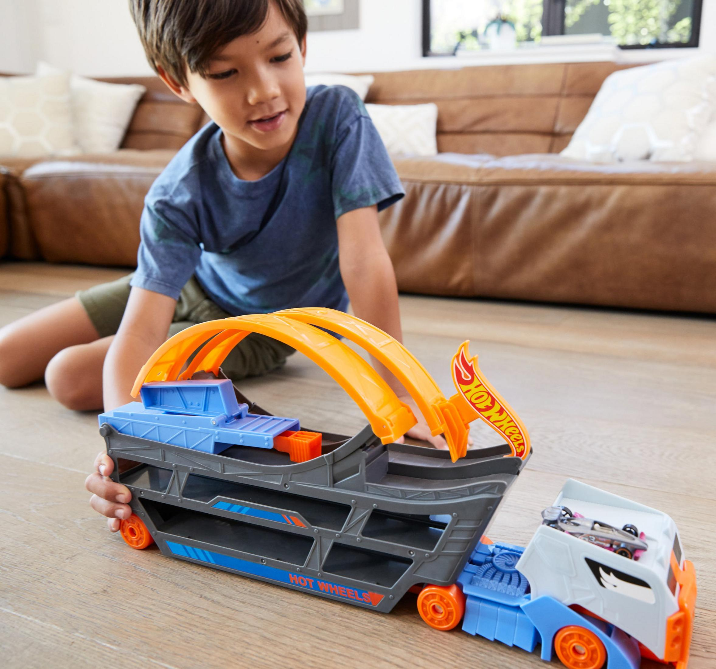 Un garçon joue avec un camion et une piste Hot Wheels sur un plancher de bois près d'un canapé.