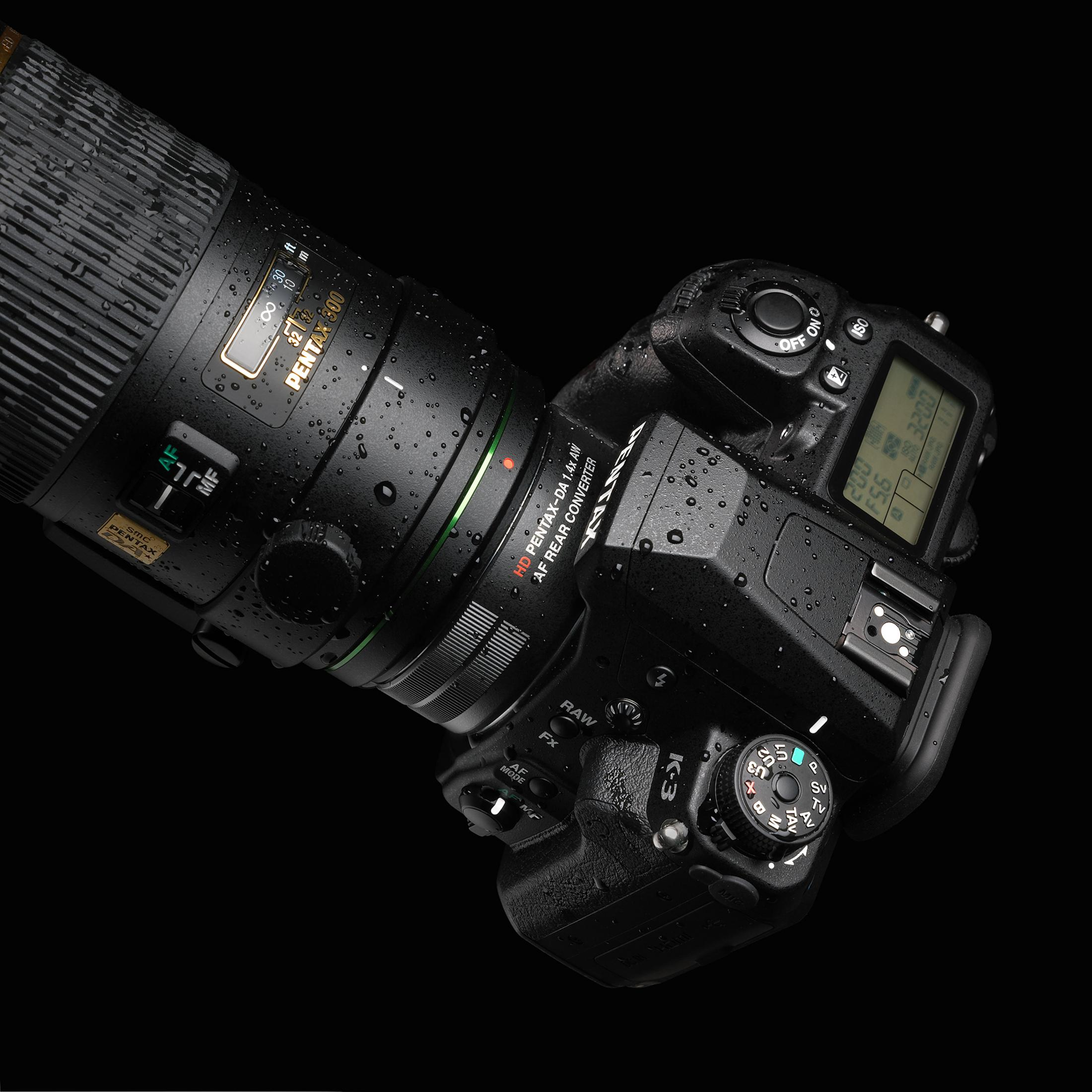 Una cámara DSLR negra con lente Pentax, cubierta de gotas de agua, sobre fondo negro.