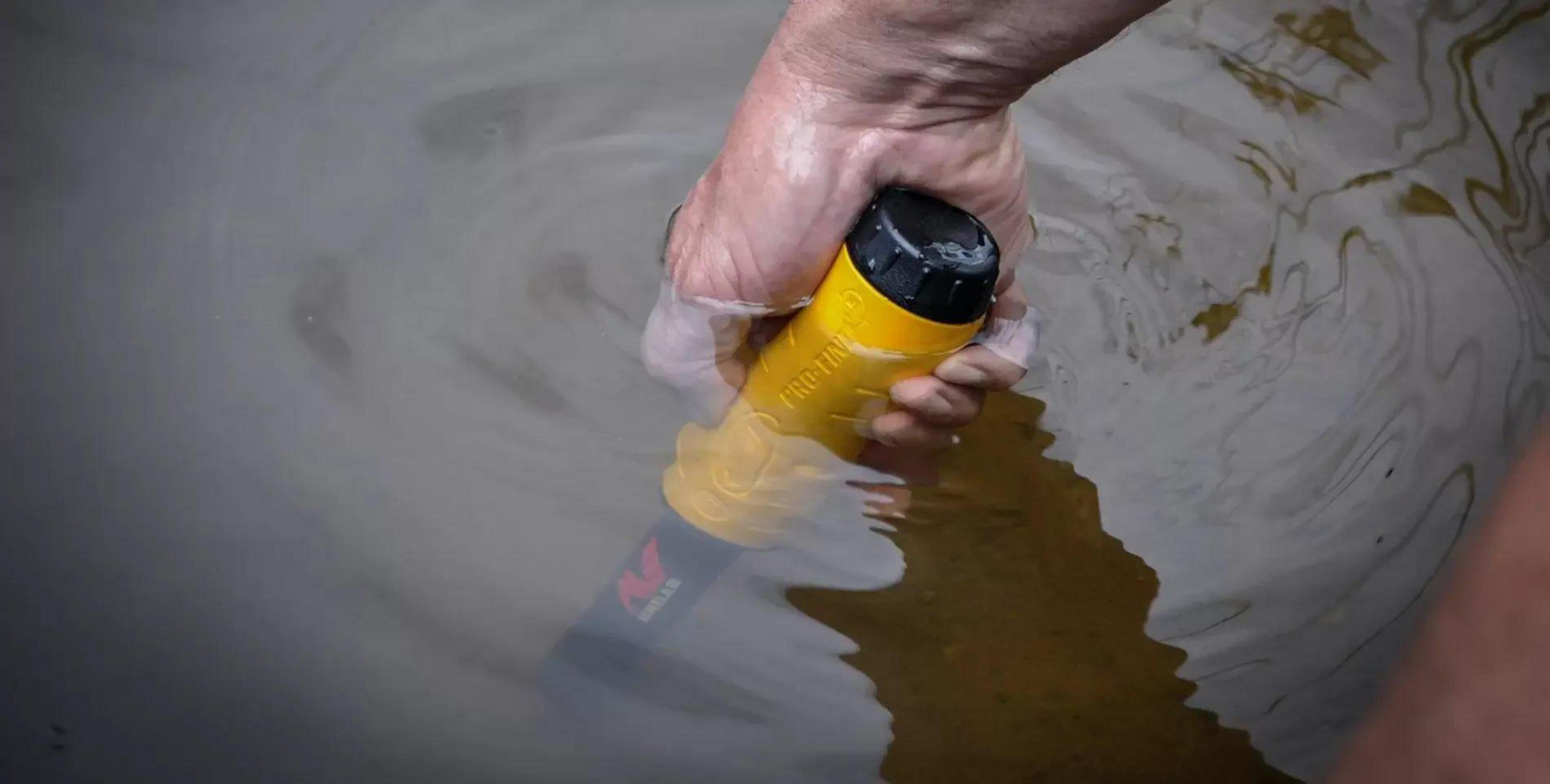 Eine Hand hält einen gelben Metalldetektor im Wasser.