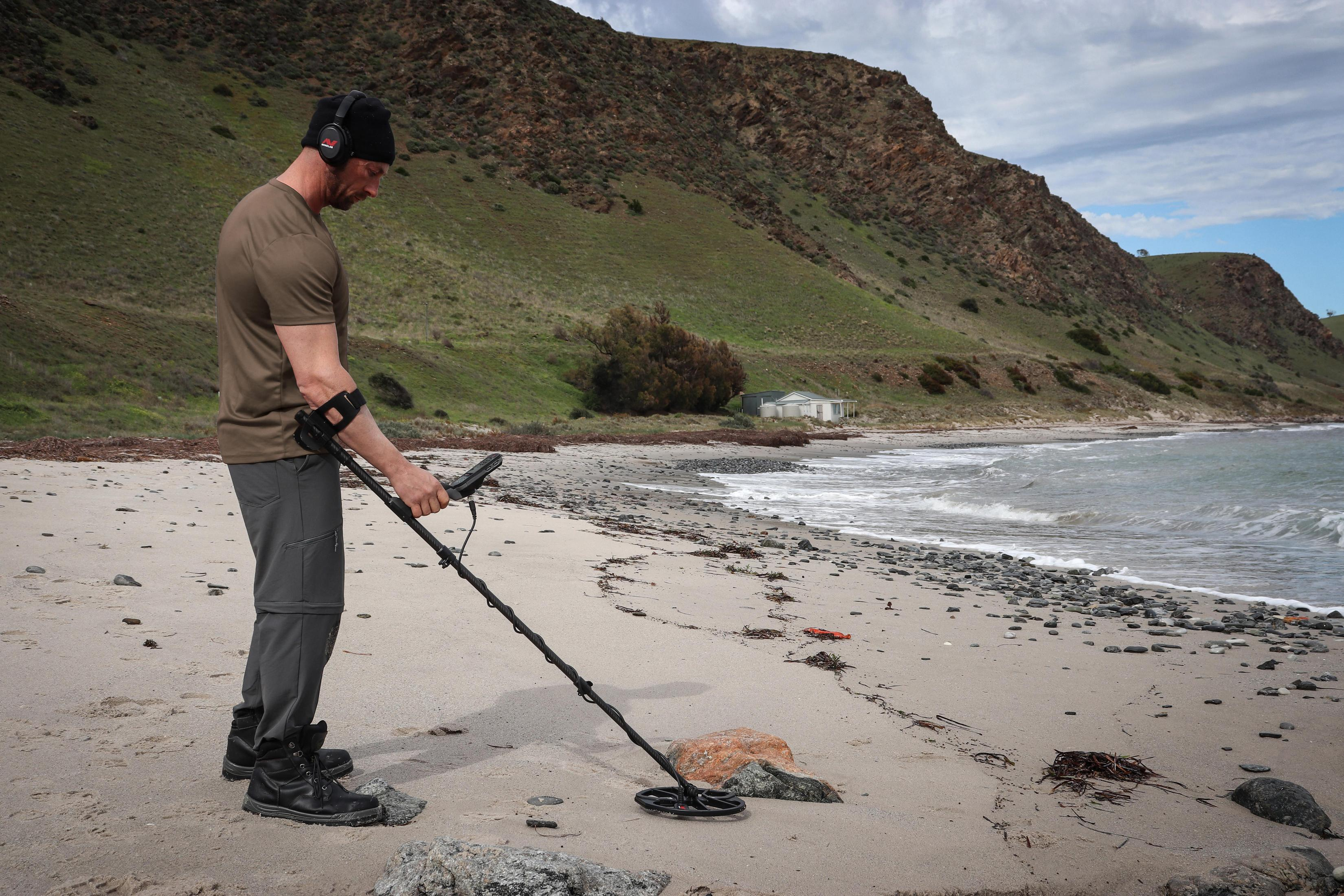Uomo con metal detector sulla spiaggia di fronte a colline e mare.