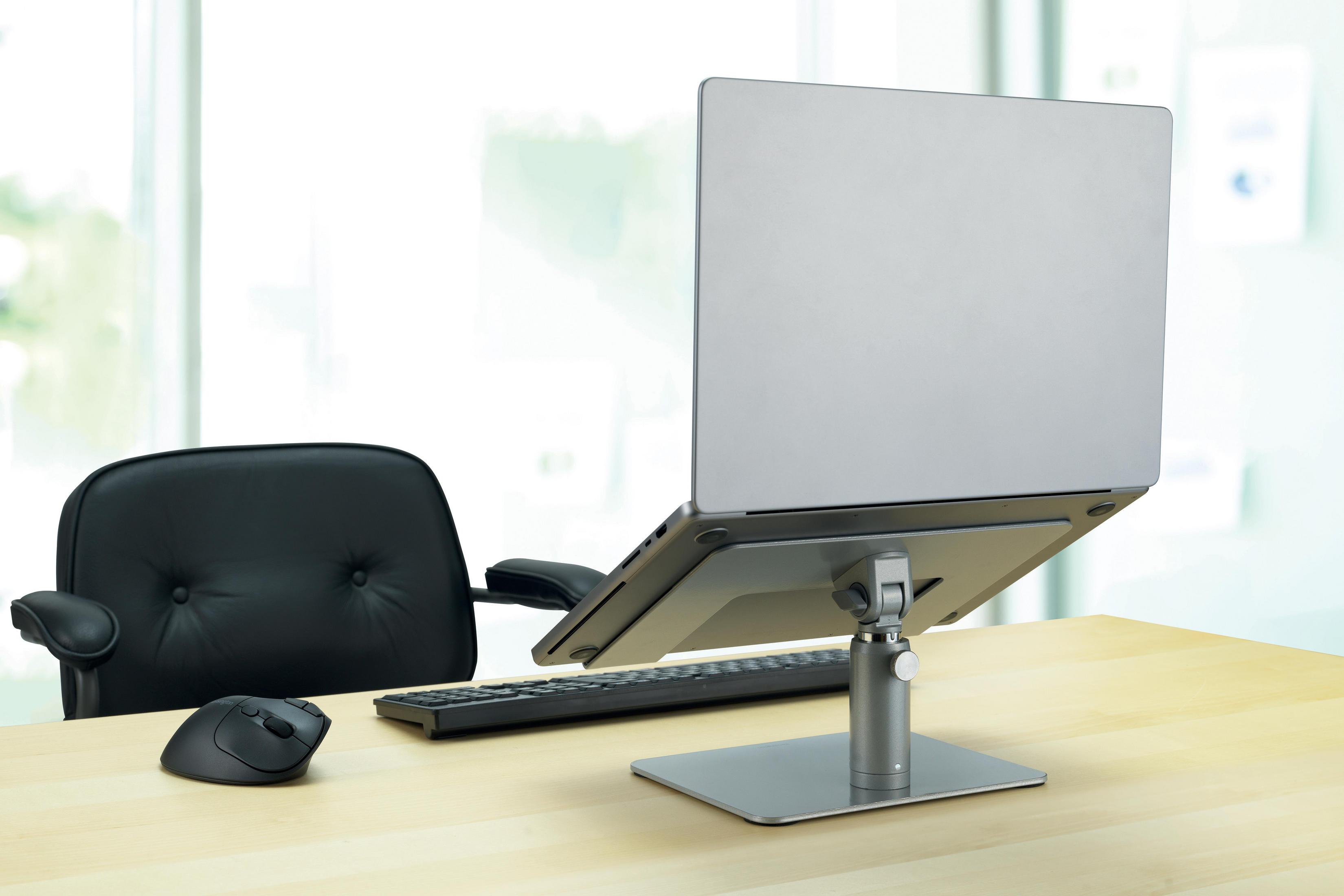 Een zilveren laptop op een standaard, met een zwart toetsenbord, muis en stoel in de buurt.