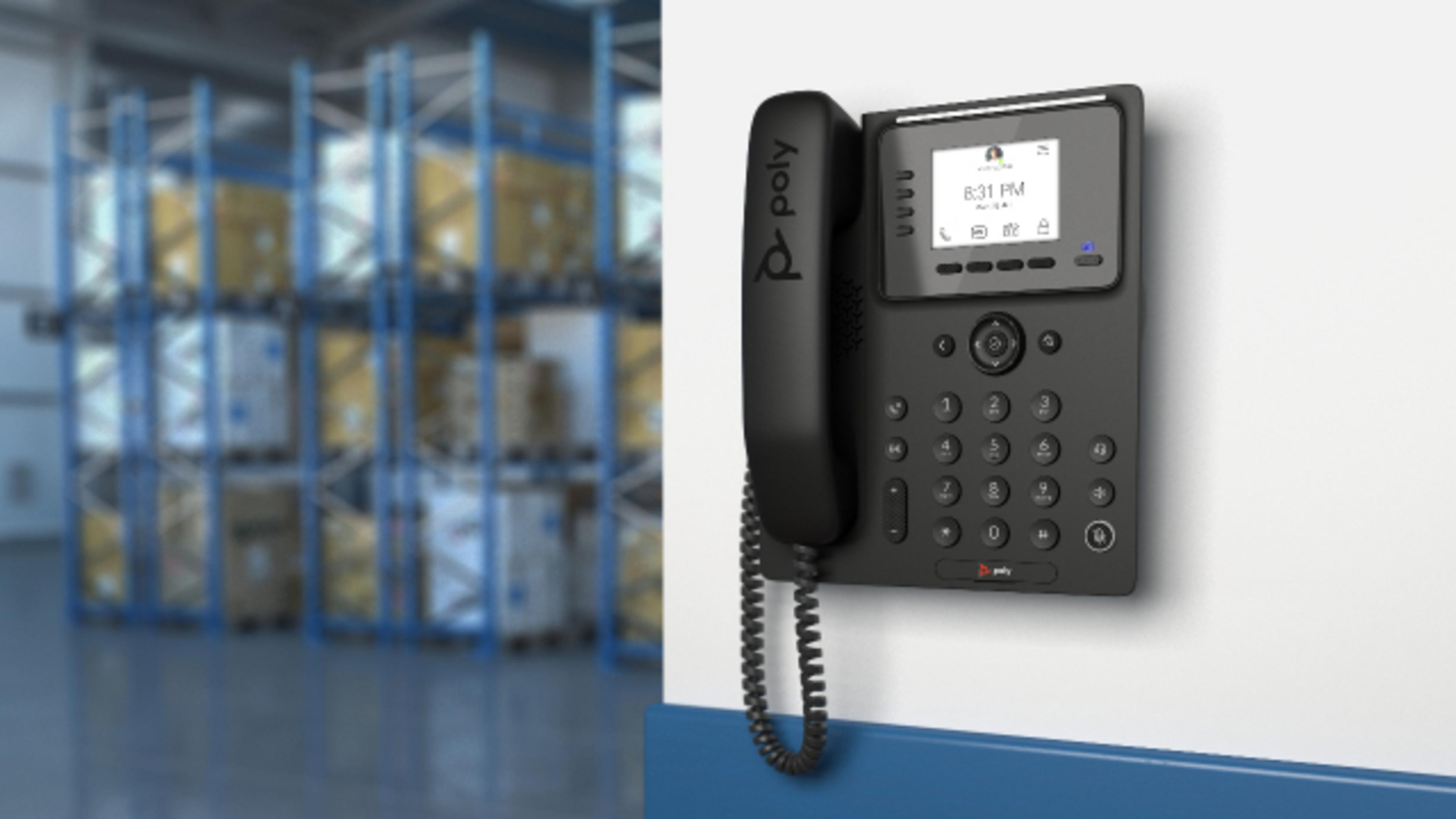 Un telefono Poly nero appeso a una parete bianca con scaffali del magazzino e scatole sullo sfondo.