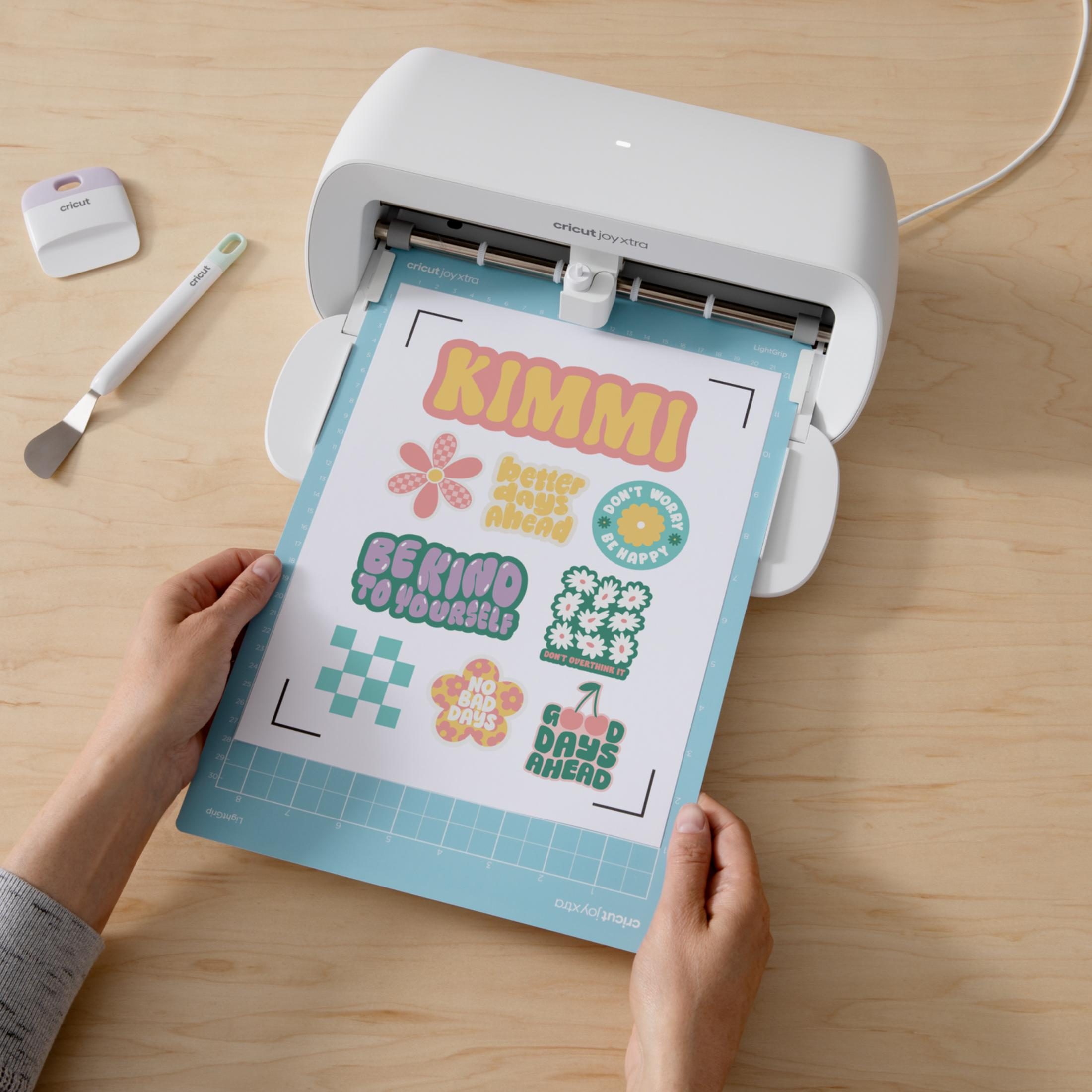 Una máquina de corte Cricut Joy Xtra con pegatinas en una alfombrilla, sostenida por manos.