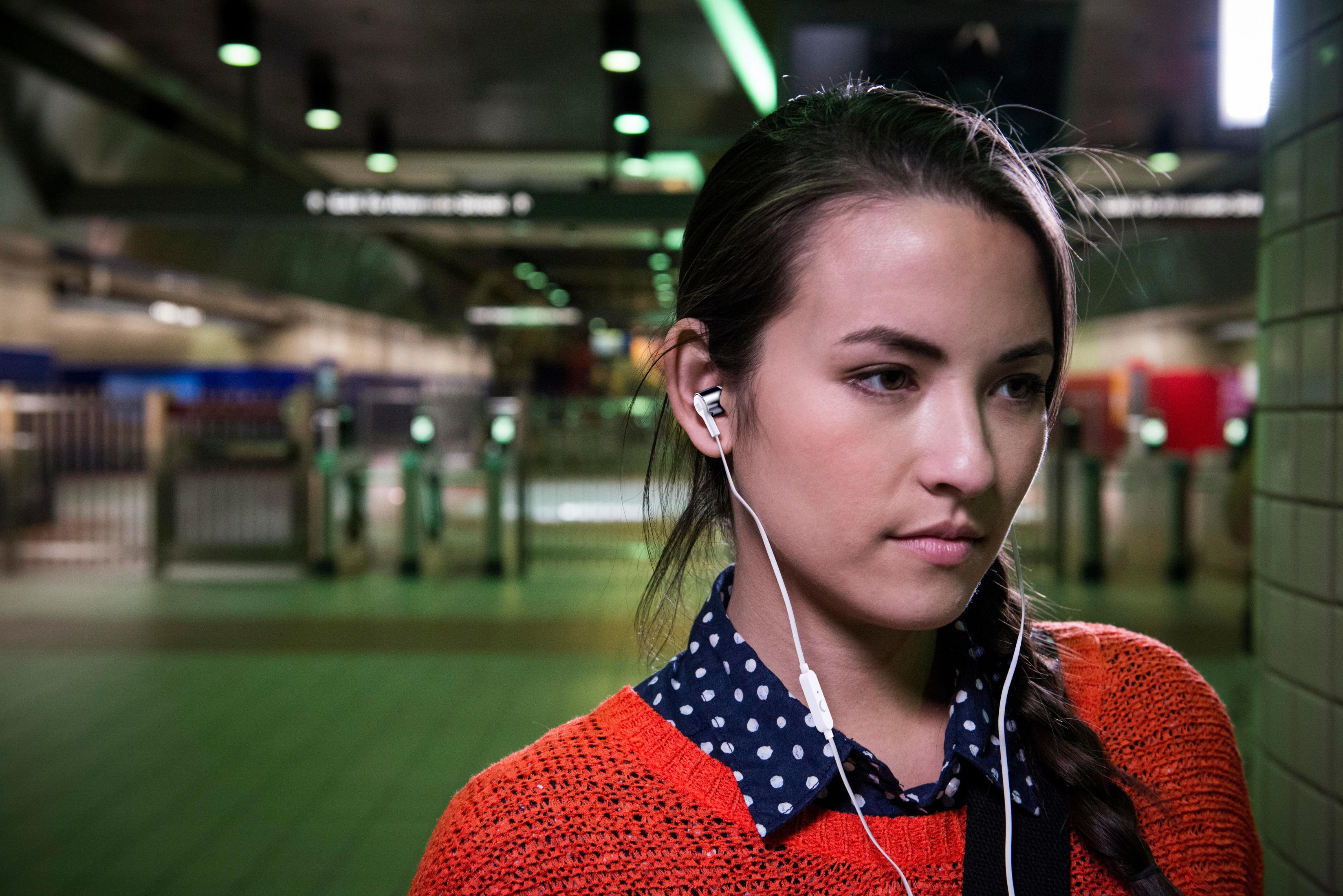Young woman with earphones in a subway station, wearing an orange sweater.