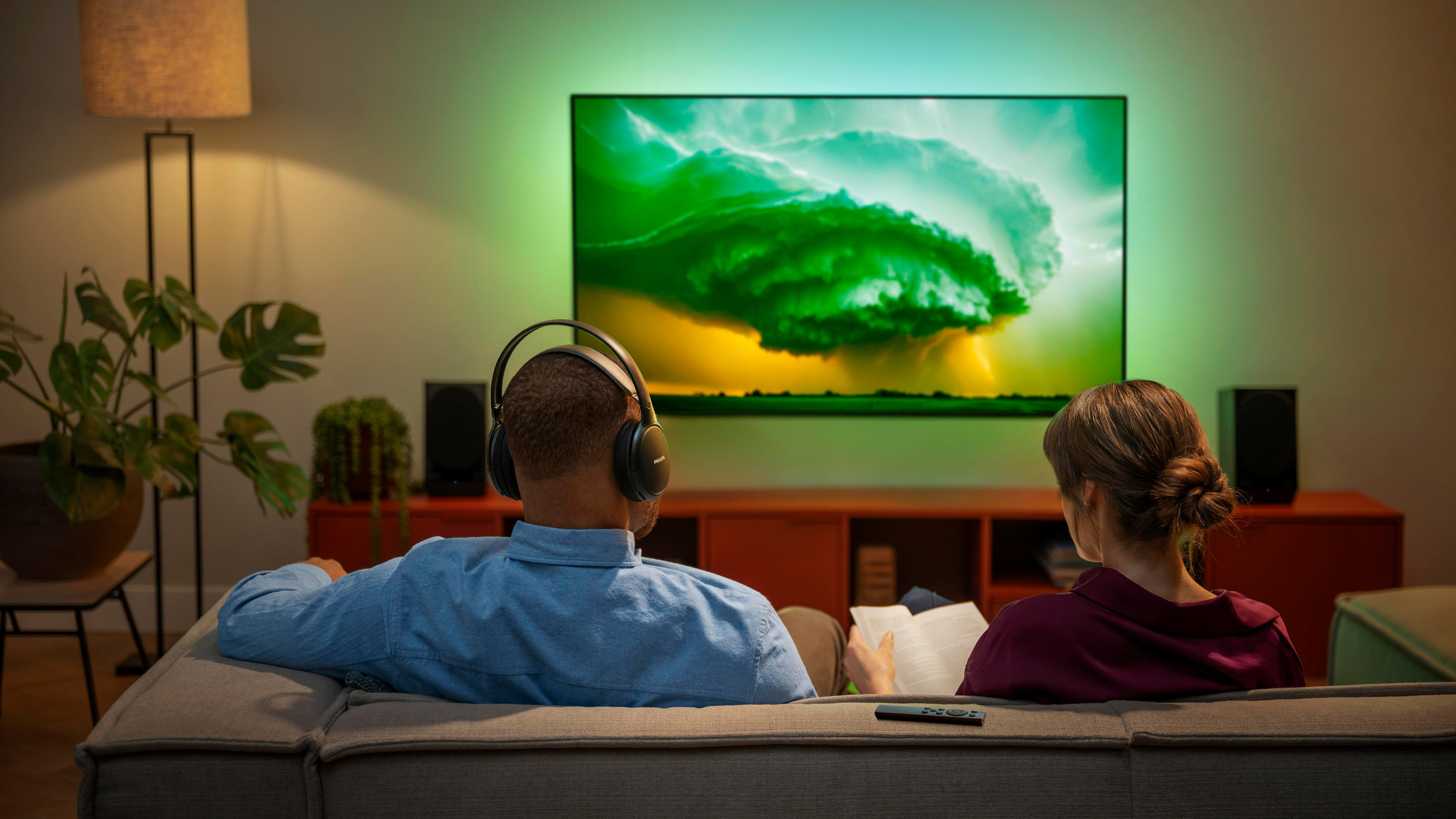 Couple watches TV, man wears headphones, woman reads book. Green ambient lighting.