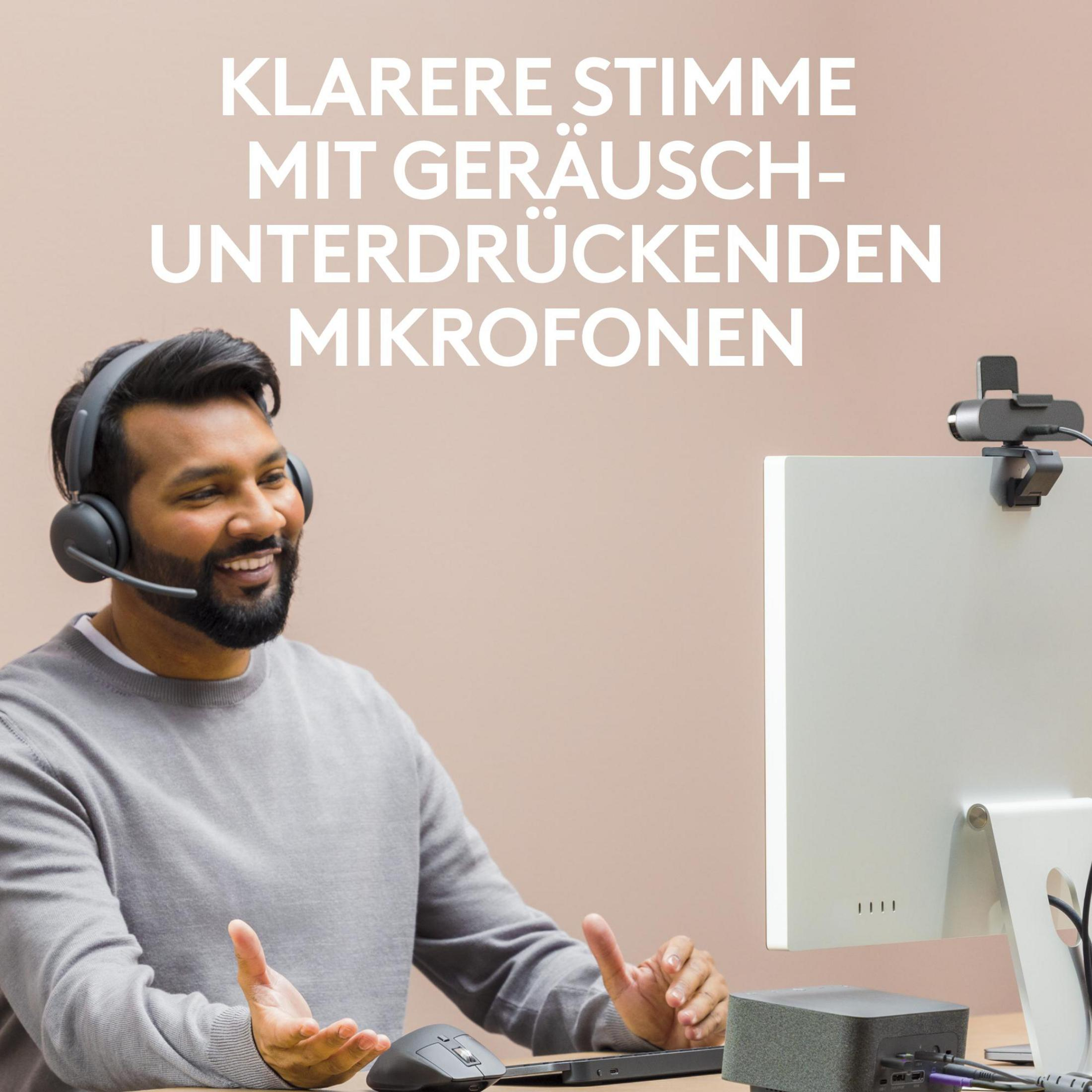 Ein Mann mit Headset lächelt, sitzt am Schreibtisch mit Computer und Webcam.