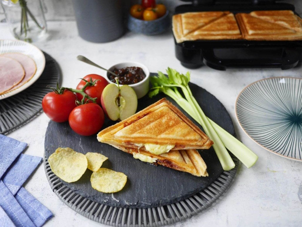 Una sandwichera con sándwiches, adornada con tomates, manzana, apio, patatas y jamón.