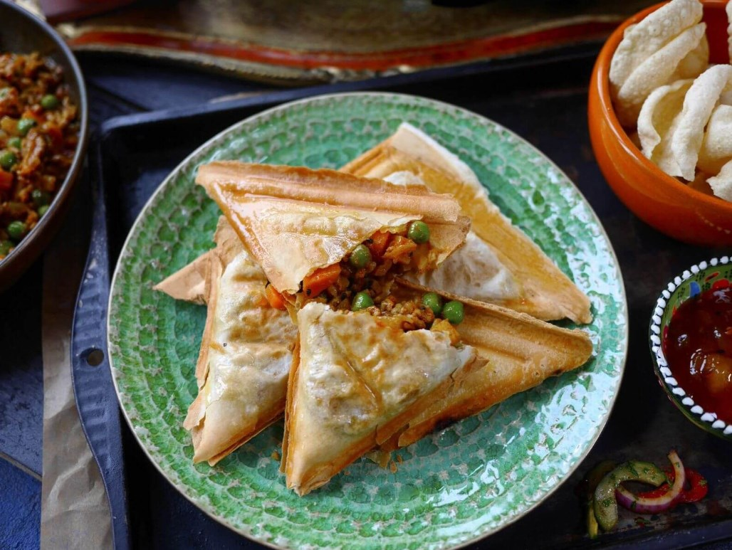 Pasteles triangulares rellenos con guisantes y arroz en un plato verde, con patatas y salsa.