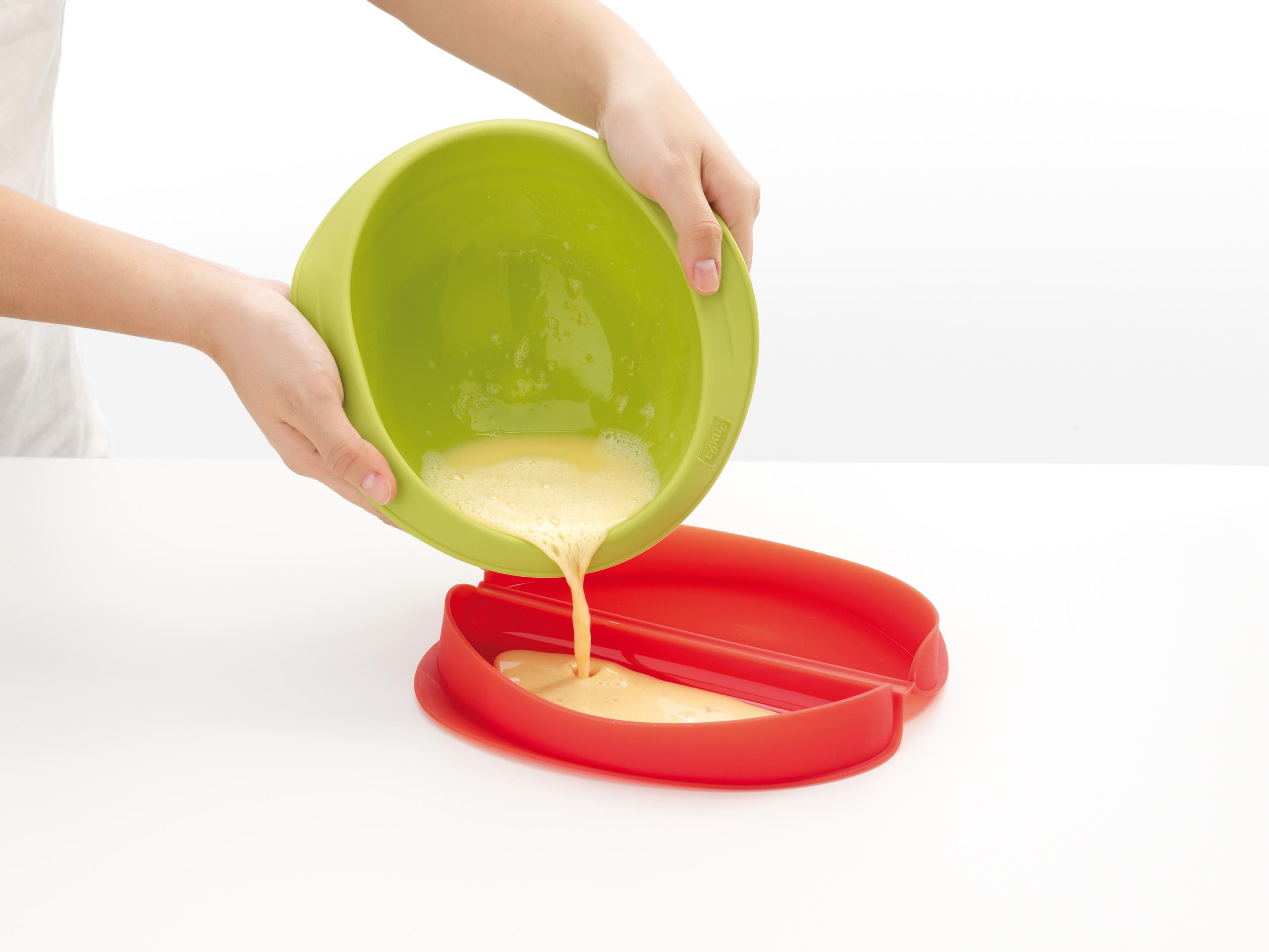 La pastella liquida viene versata da una ciotola verde in uno stampo rosso.