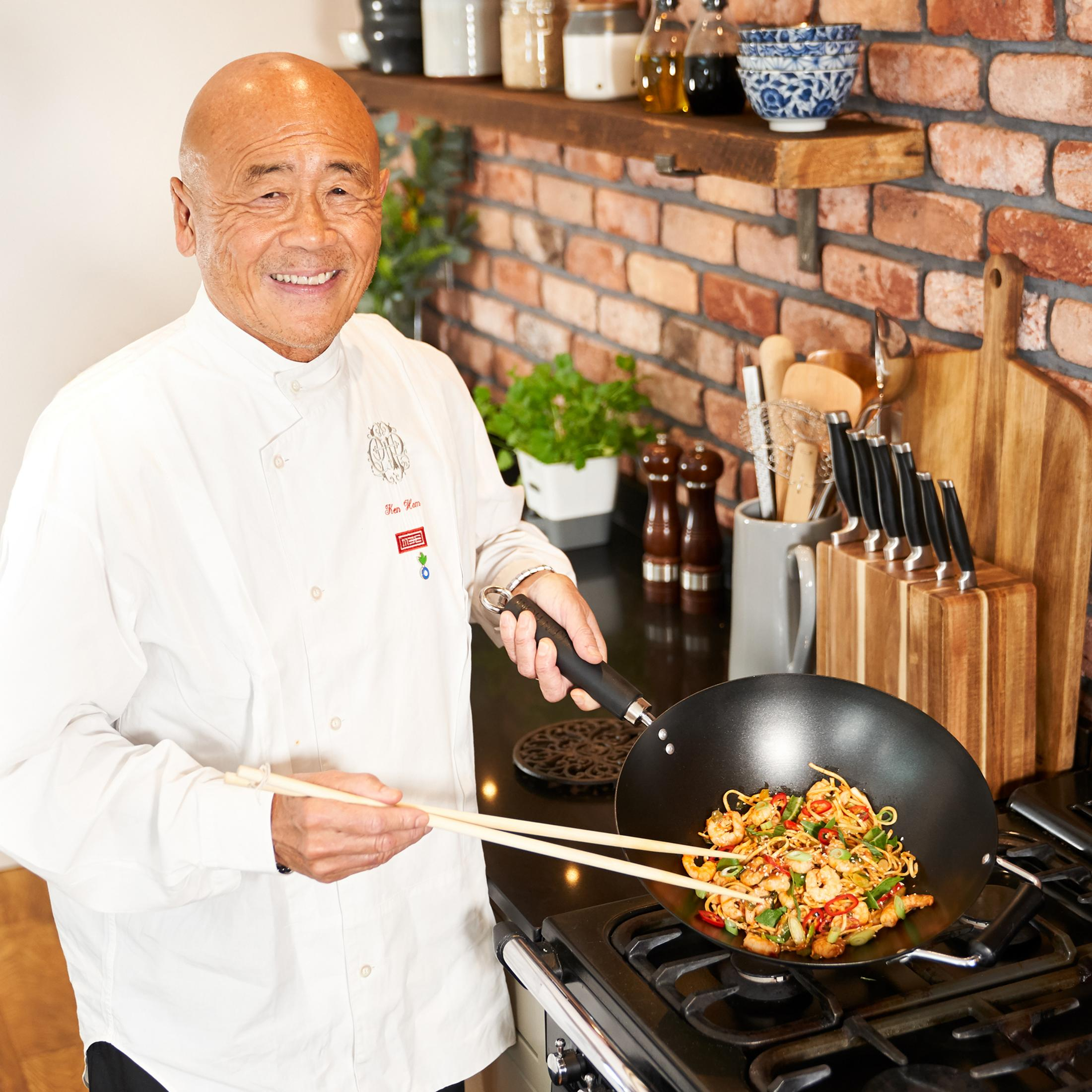Un chef en blanc cuisine des nouilles aux crevettes dans un wok sur une cuisinière à gaz.
