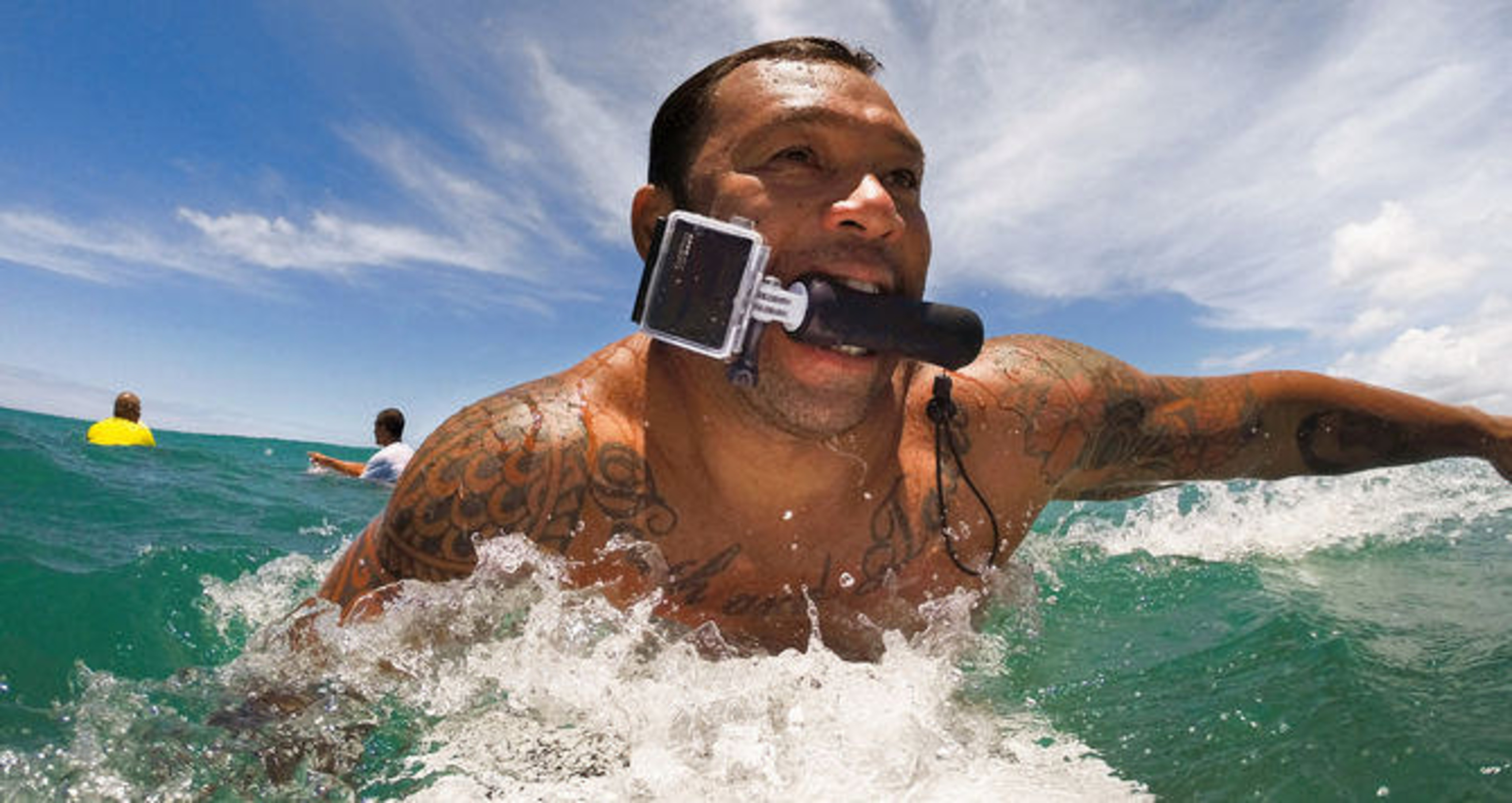 Mann mit Tattoo im Wasser, Kamera im Mund, surft auf einer Welle.