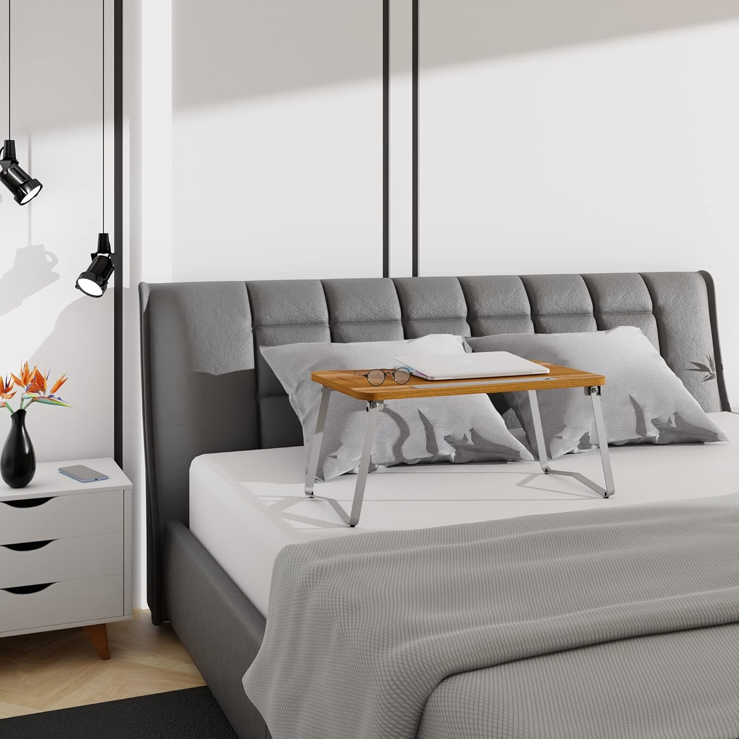 Una bandeja de madera para portátil con un portátil y gafas en una cama con ropa de cama gris.