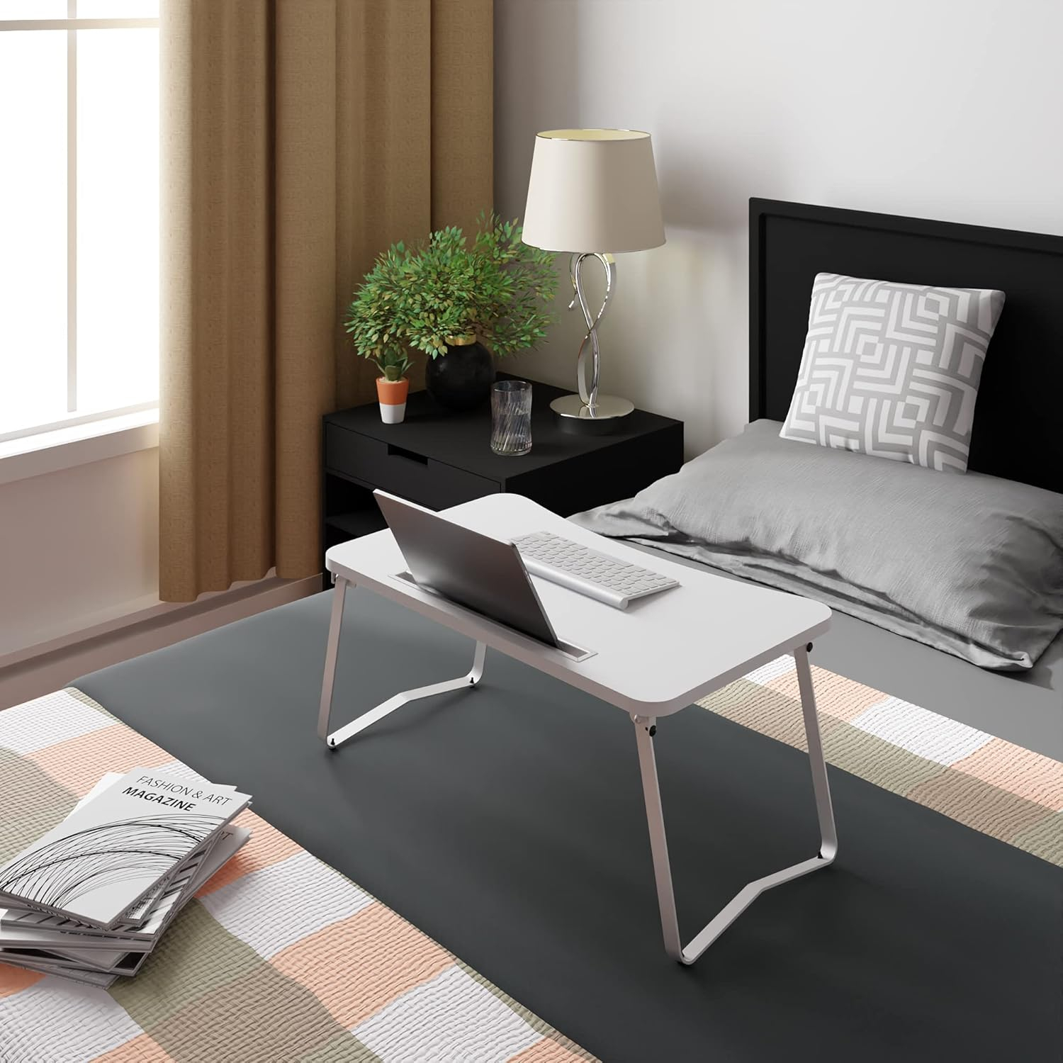 Una mesa blanca para portátil con portátil está sobre una cama junto a una mesita de noche con lámpara.