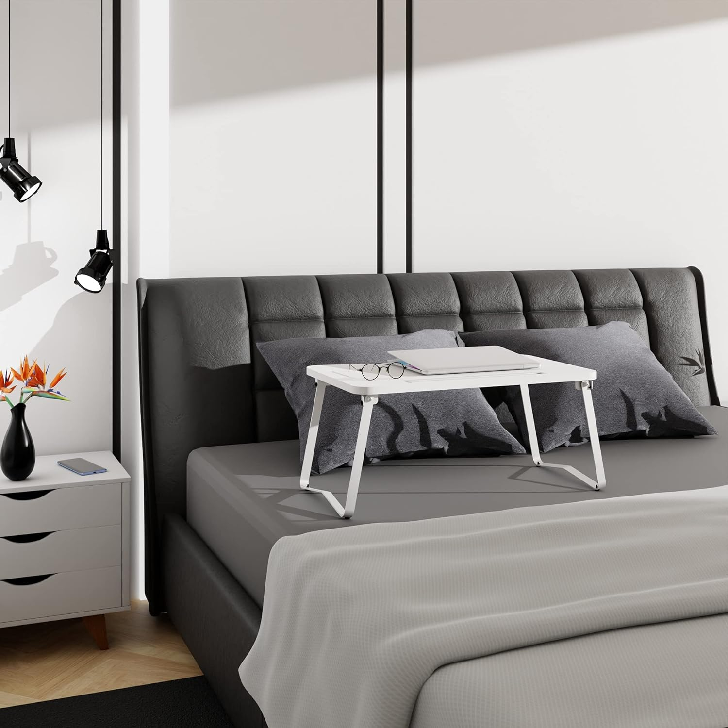 Una mesa blanca para portátil está sobre una cama con almohadas grises. Una mesita de noche al lado.