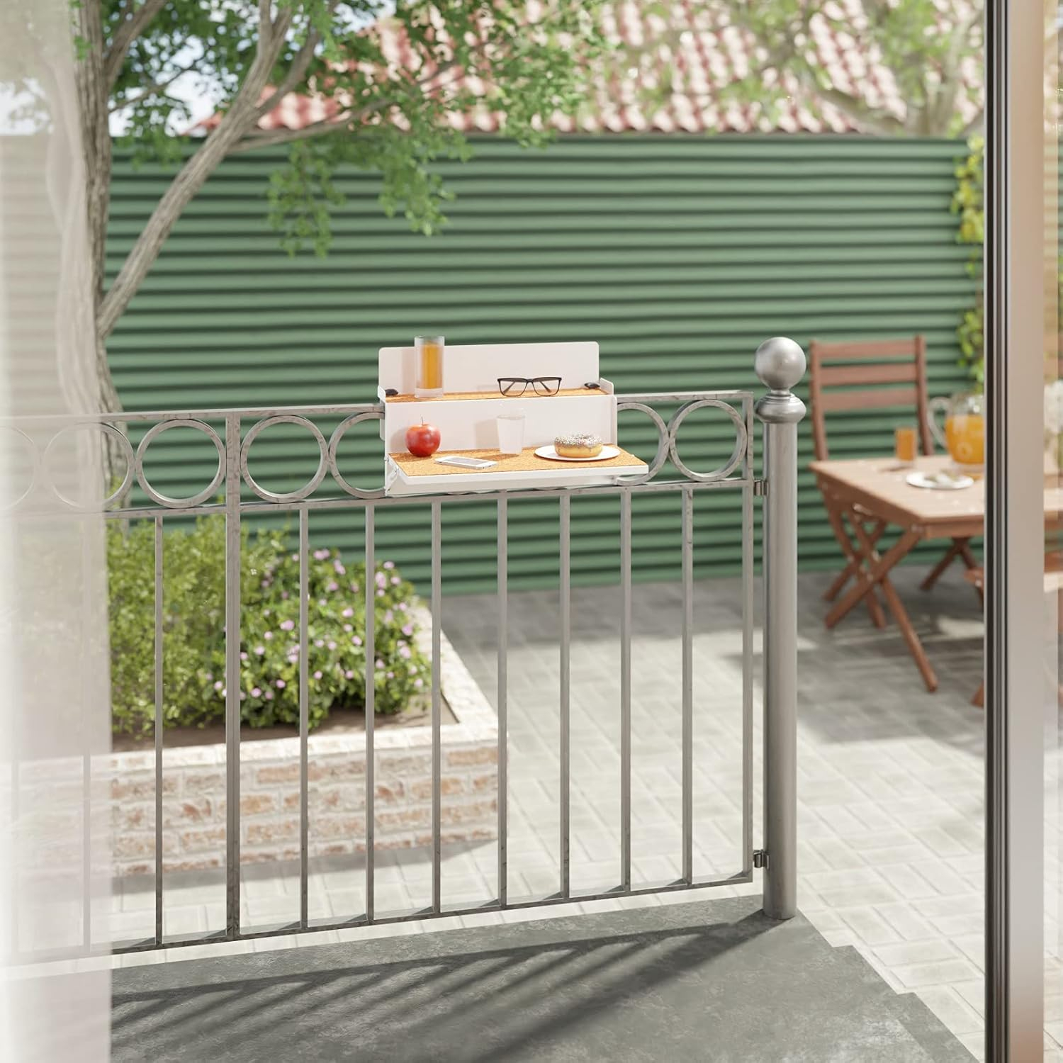 Una mesa de balcón blanca con una manzana roja, vasos y una rosquilla está unida a una barandilla gris con vistas a un jardín verde.