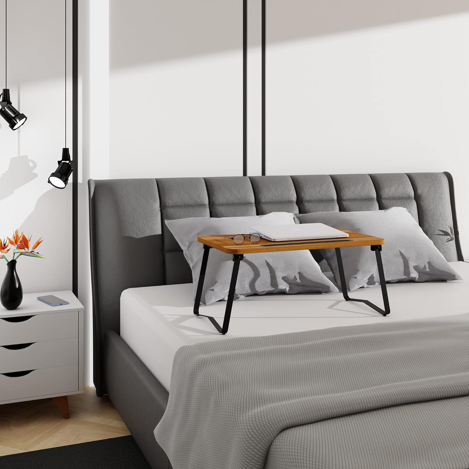 Un portátil y gafas en una bandeja sobre una cama con almohadas grises.