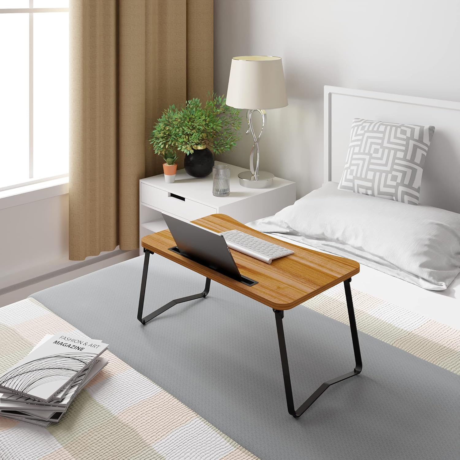 Una mesa de madera para portátil con portátil y teclado sobre una cama. Al lado hay una mesita de noche con lámpara y planta.