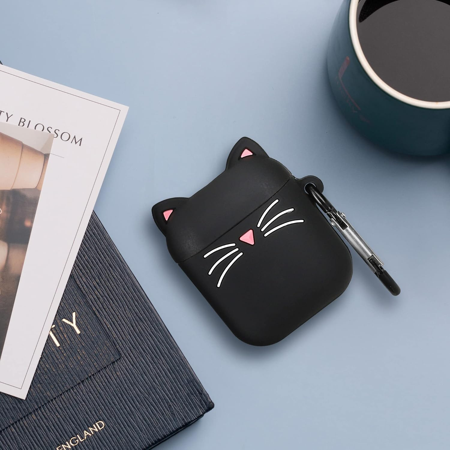 Schwarzer Katzen-Kopfhörerhülle mit rosa Ohren und Schnurrhaaren, neben einem Buch und einer Tasse.