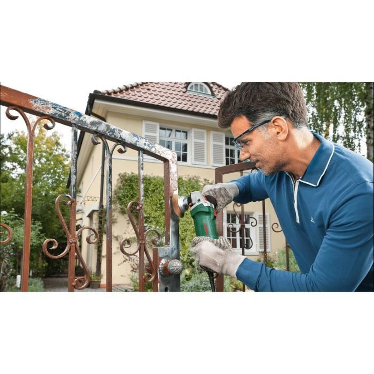 Mann in blauem Hemd und Schutzbrille schleift rostiges Metalltor mit Elektrowerkzeug.