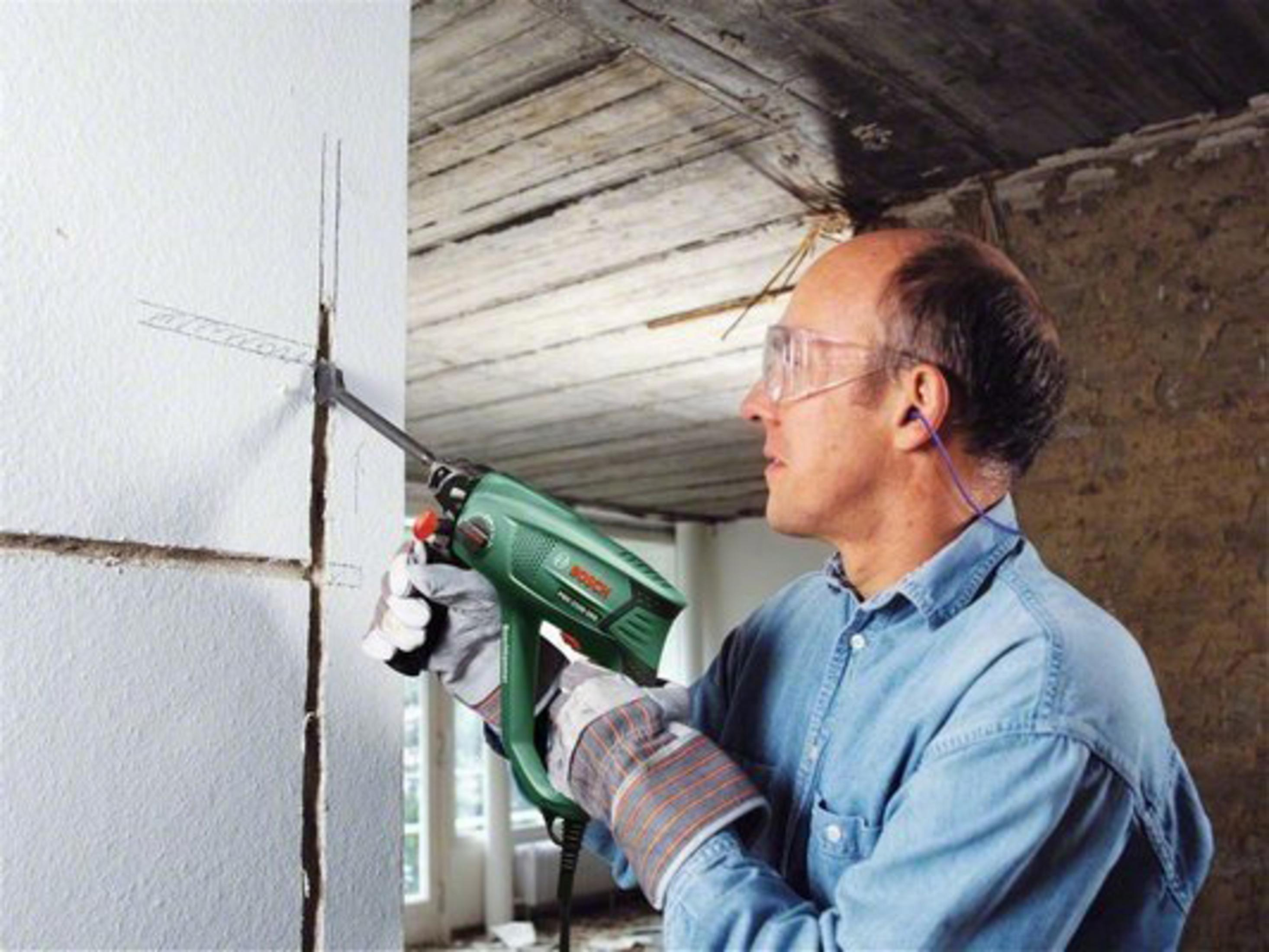 Mann mit Schutzbrille und Gehörschutz benutzt Bohrmaschine an Wand.