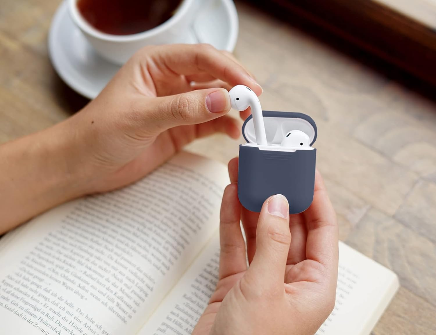 Hände halten AirPods Ladecase mit Ohrhörern vor einem Buch und Tasse Tee.