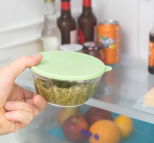 Mano tiene ciotola trasparente con cibo verde e coperchio verde chiaro in frigo.