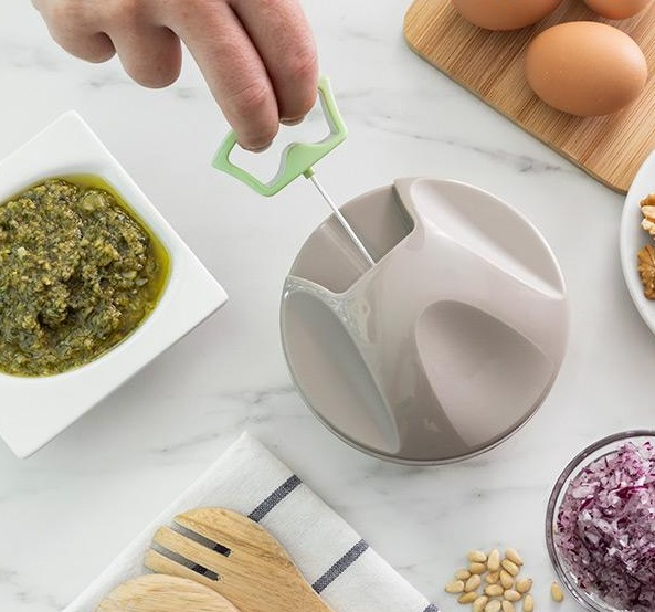 Mano che usa un gadget da cucina, accanto una ciotola di pesto, uova e cipolle.