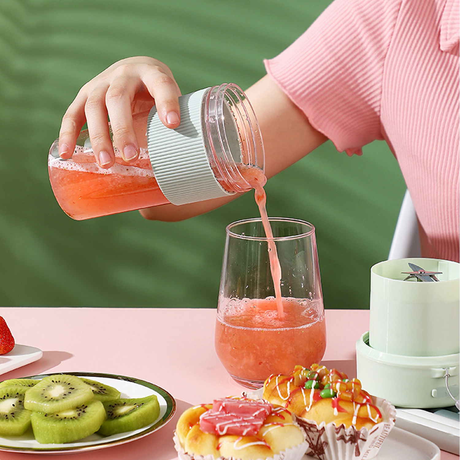 Una mano versa succo rosso da un frullatore portatile in un bicchiere. Kiwi e pasticcini su un tavolo.