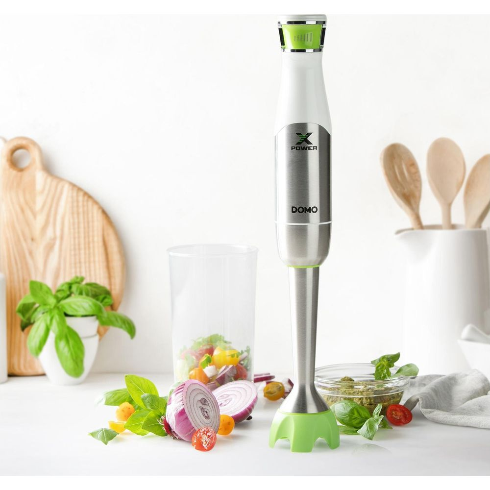 Ein Stabmixer steht auf einer Arbeitsplatte mit Zutaten wie Zwiebeln, Tomaten und Kräutern.