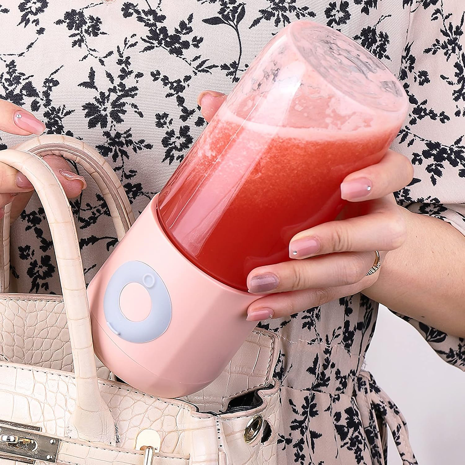Una persona tiene un frullatore portatile rosa con succo rosso vicino a una borsa beige.