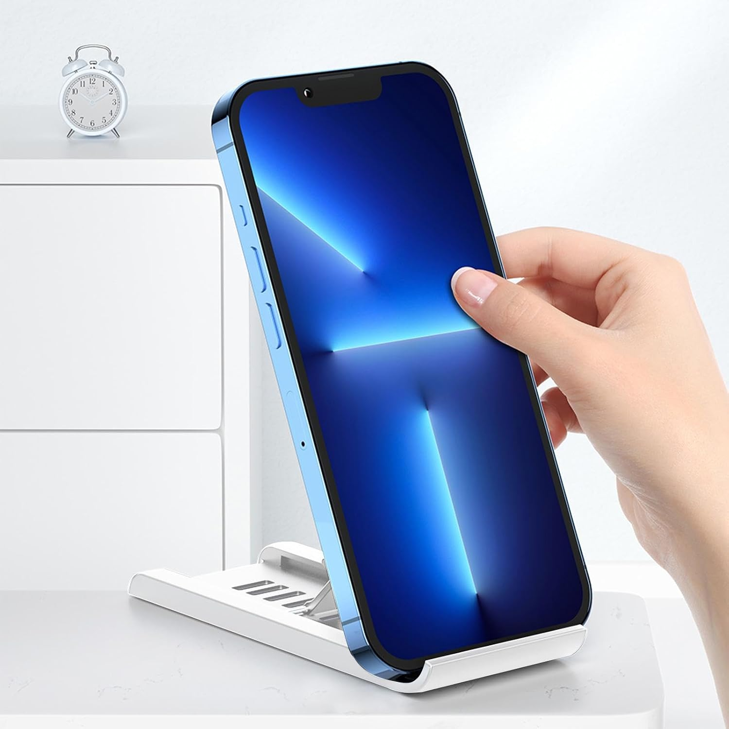 Eine Hand hält ein blaues Smartphone auf einem weißen Ständer. Ein kleiner Wecker steht links.