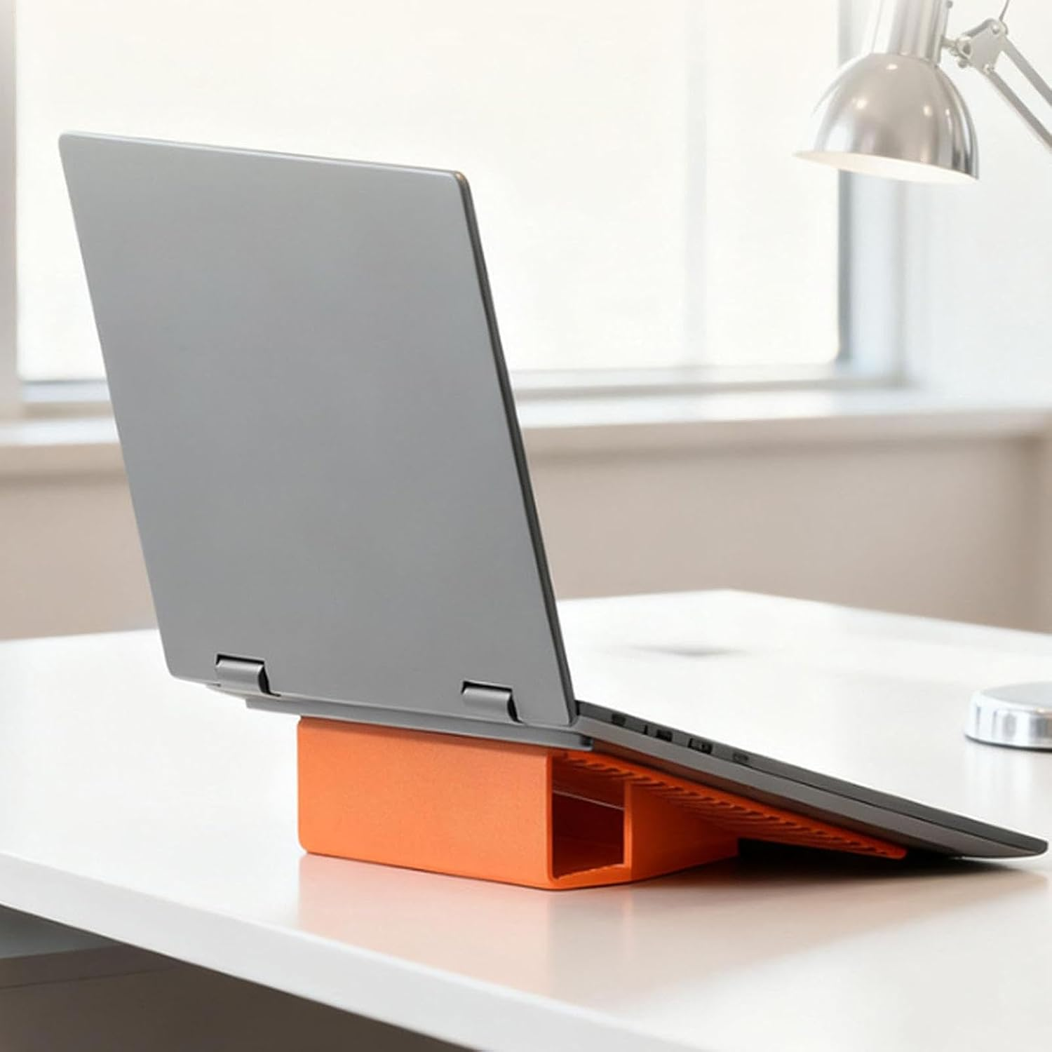 Ein graues Laptop steht auf einem orangefarbenen Ständer auf einem weißen Schreibtisch.