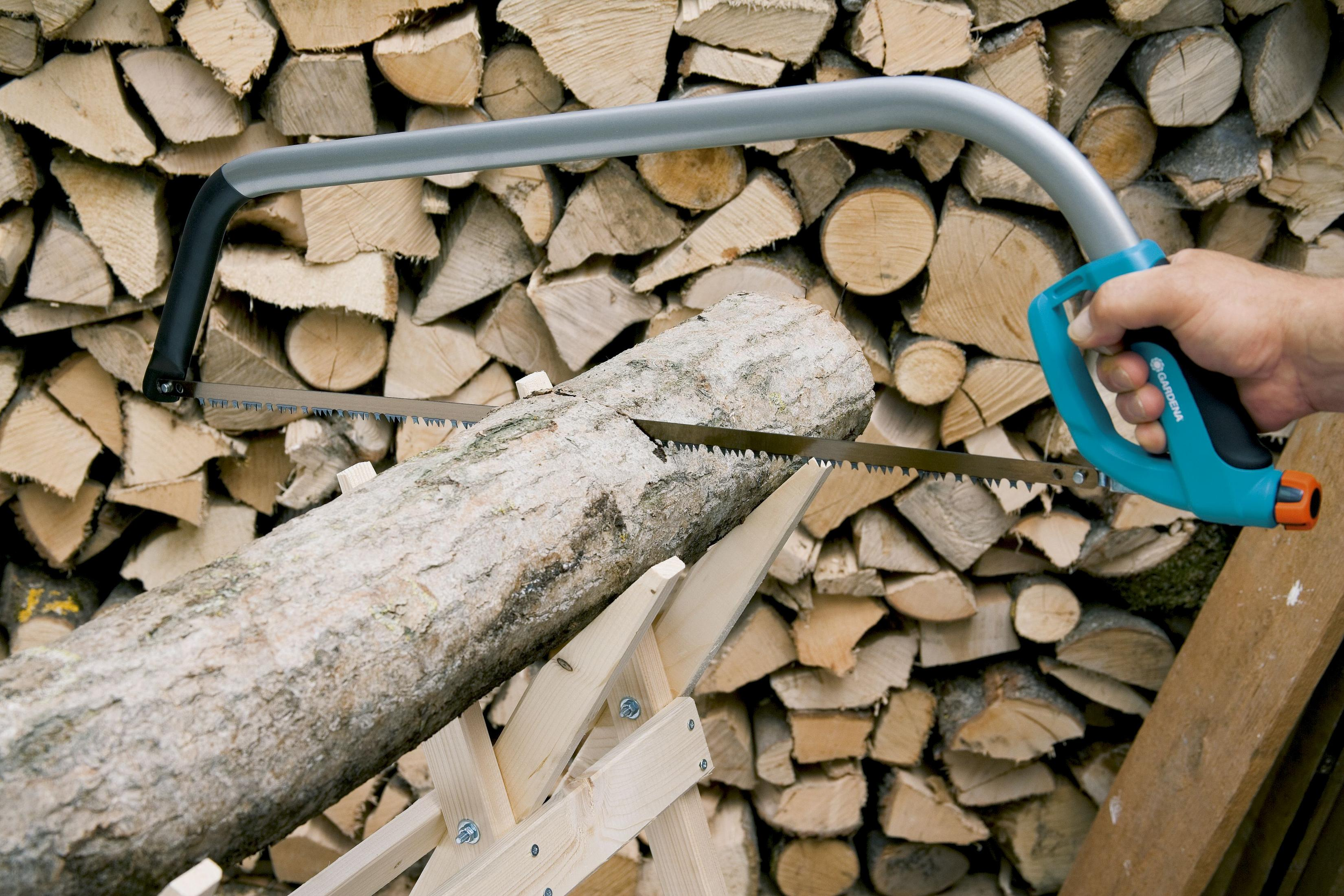 Ein Mann sägt mit einer Bügelsäge einen Baumstamm, der auf einem Holzgestell liegt. Im Hintergrund liegt aufgeschichtetes Brennholz.