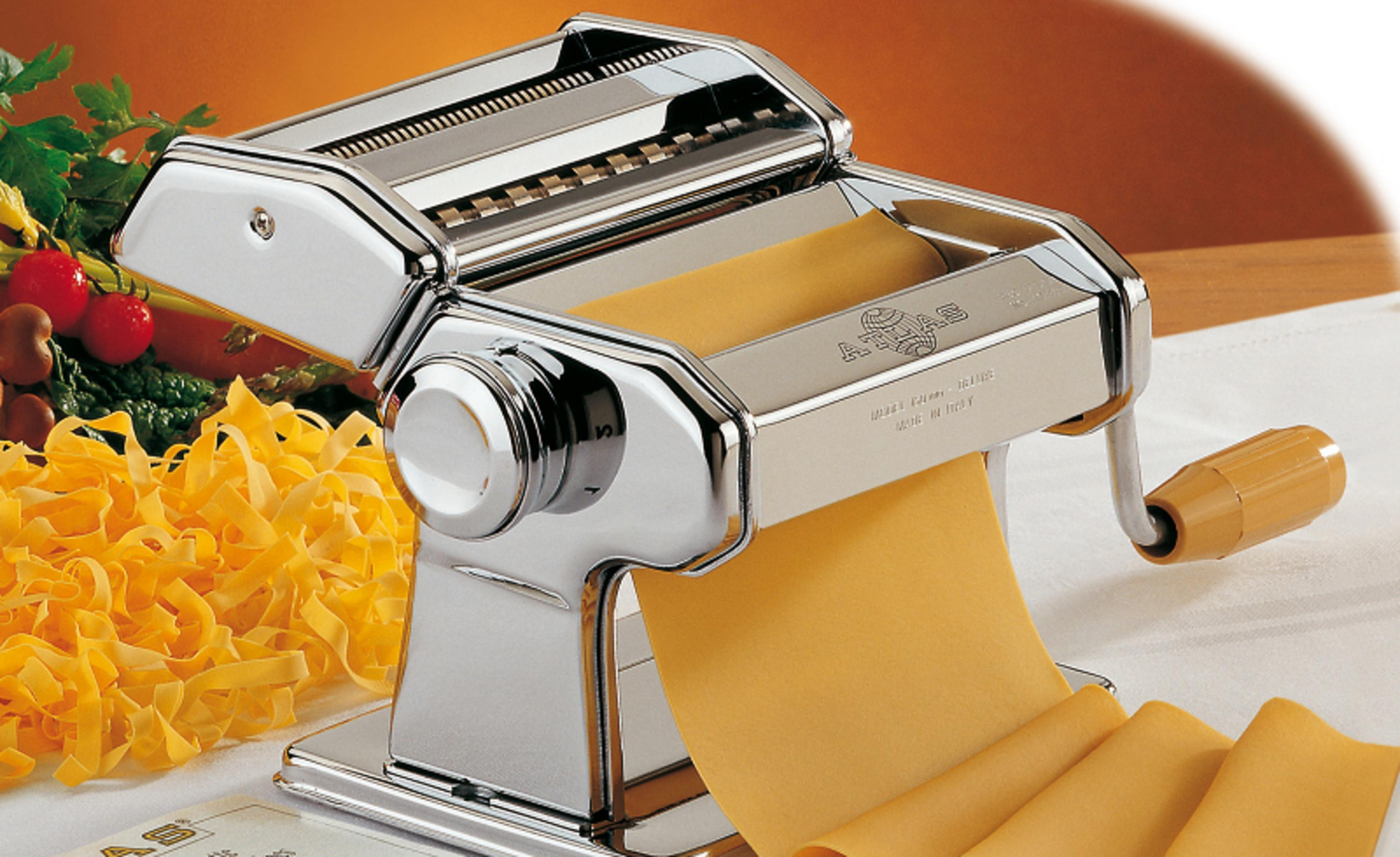 Macchina per pasta cromata lucida con impasto giallo, accanto a pasta fresca e pomodori.