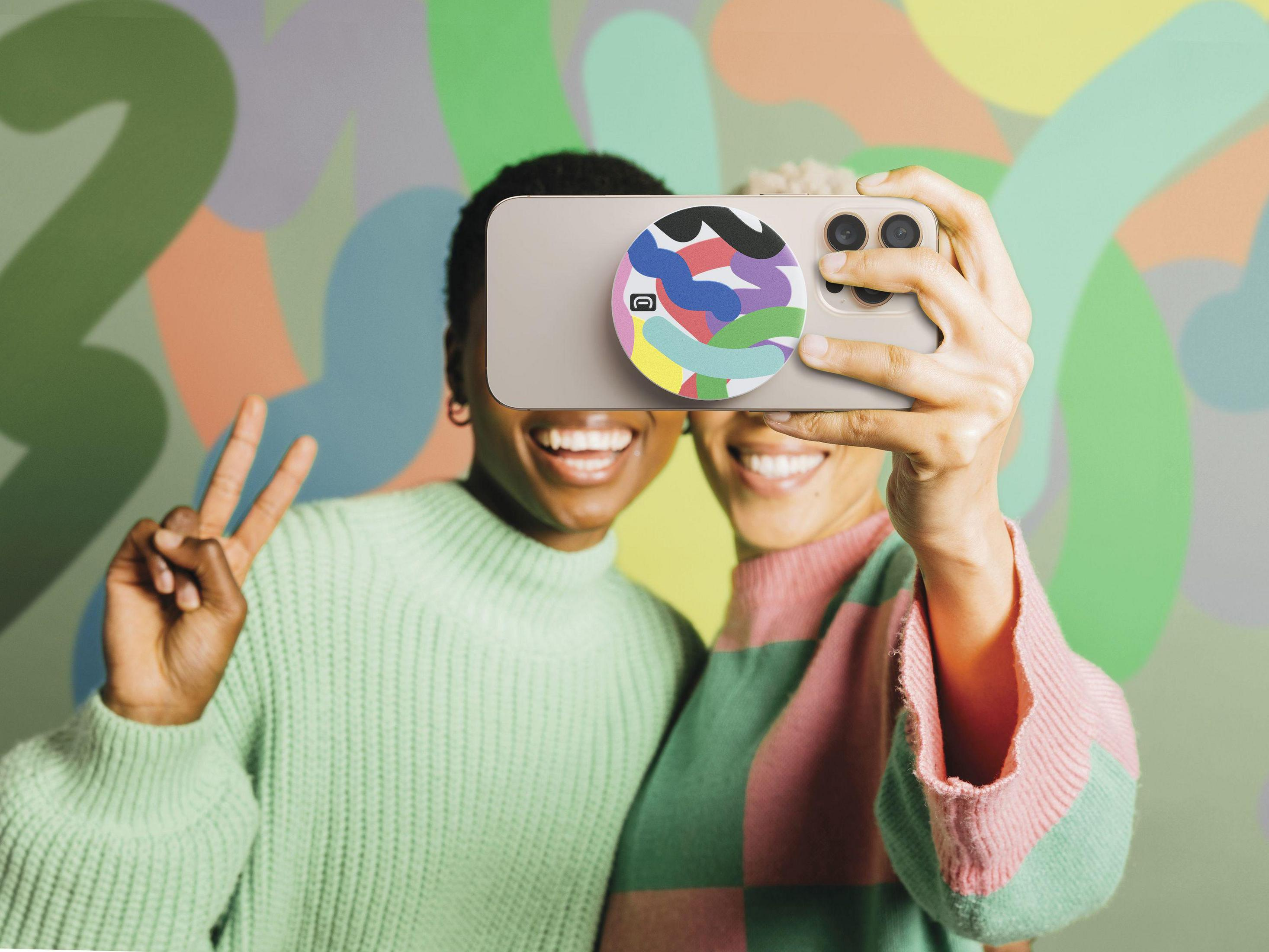 Due donne si scattano un selfie davanti a un muro colorato. Una fa il segno della pace.