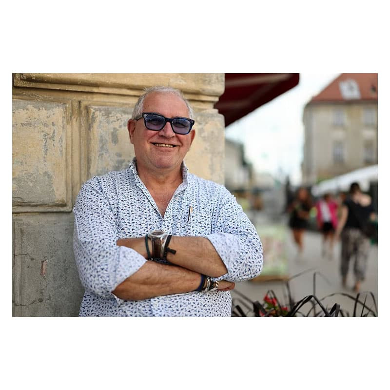 Un hombre con gafas de sol y camisa estampada está de pie con los brazos cruzados junto a un muro.